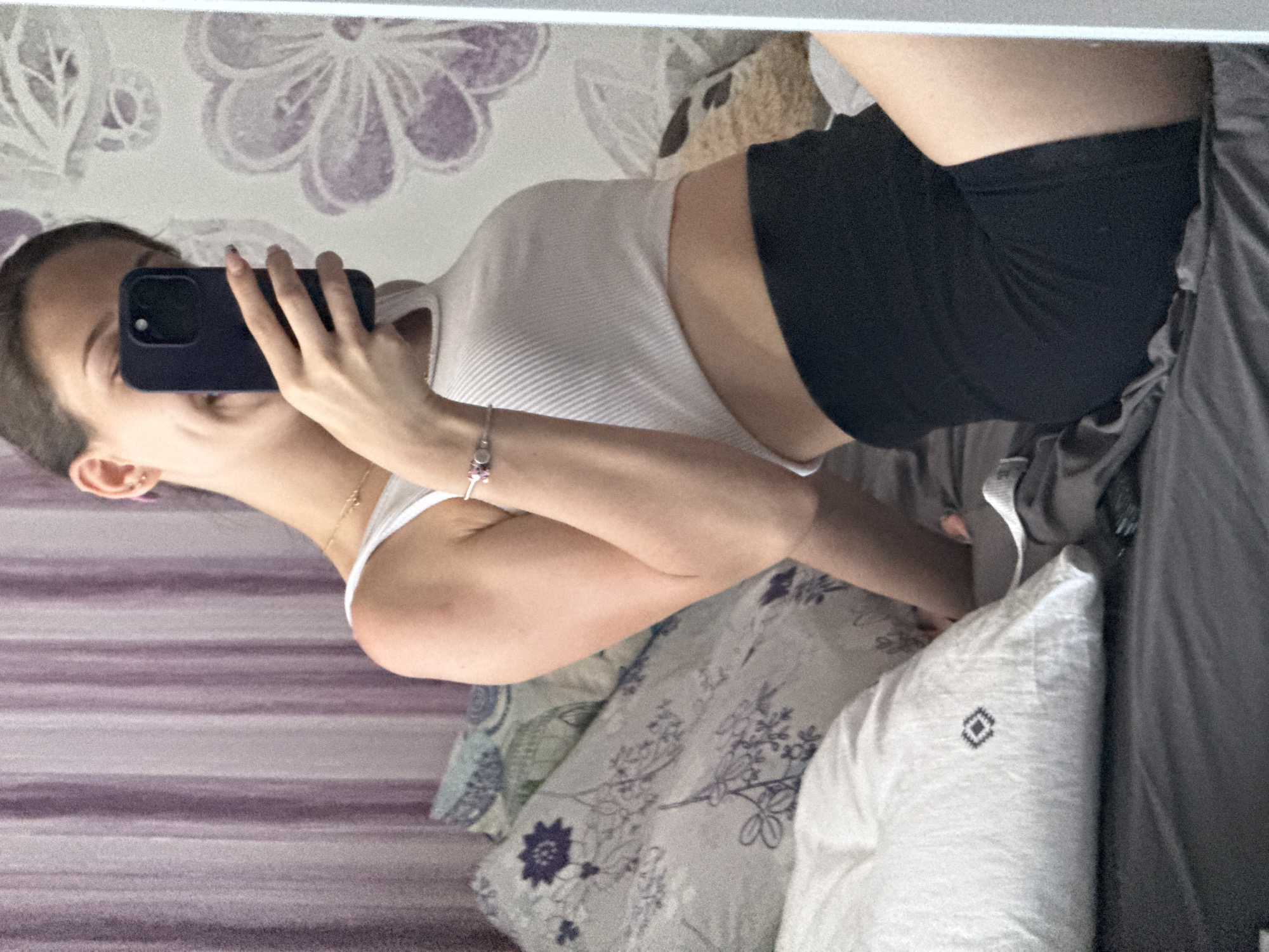A young woman, seen from the waist up in a mirror reflection, is taking a selfie. She is wearing a white ribbed crop top and black biker shorts. She has a pink stud earring in her left ear and a bracelet on her left wrist with pink charms. She is holding a dark blue smartphone in her right hand, obscuring part of her face. The background appears to be a bedroom with a patterned purple and white floral wallpaper. There are pillows on a bed, one of which is white with a dark diamond pattern. The scene is indoors, with soft lighting, and suggests a casual, personal moment. The image does not provide any information that would allow for determining the location in Sofia, Bulgaria.