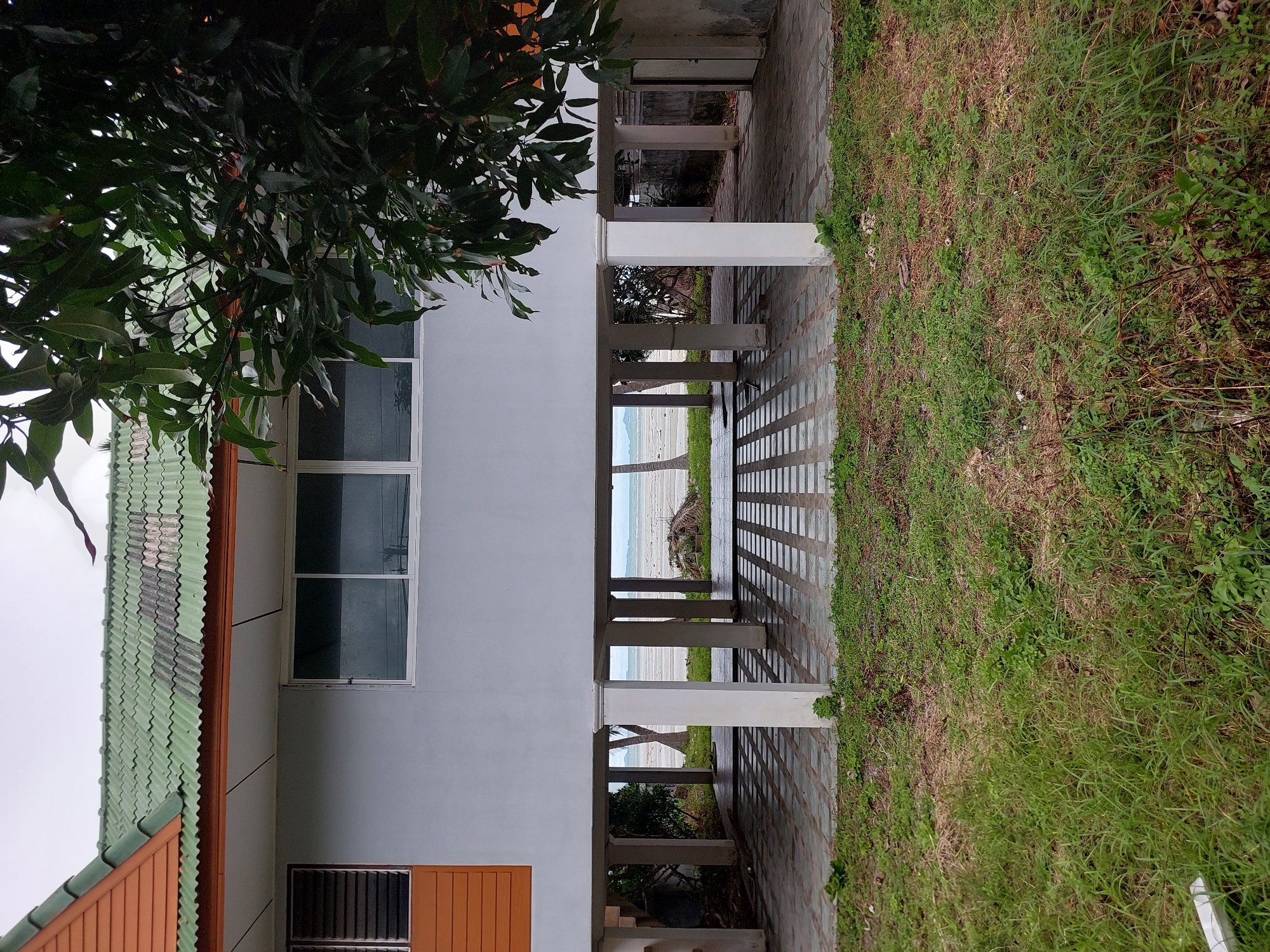 The image depicts a house raised on stilts in Ko Samui District, Thailand. The house has a green tiled roof and white walls. Large windows with white frames are visible on the upper floor. The ground floor, open beneath the raised structure, is paved with square tiles and supported by white pillars. Beyond the house, a beach and the sea are visible under an overcast sky. Vegetation, including a large tree with dark green leaves, partially obscures the view of the house. The foreground consists of a patch of overgrown grass and some dry foliage. There are no people visible in the image.