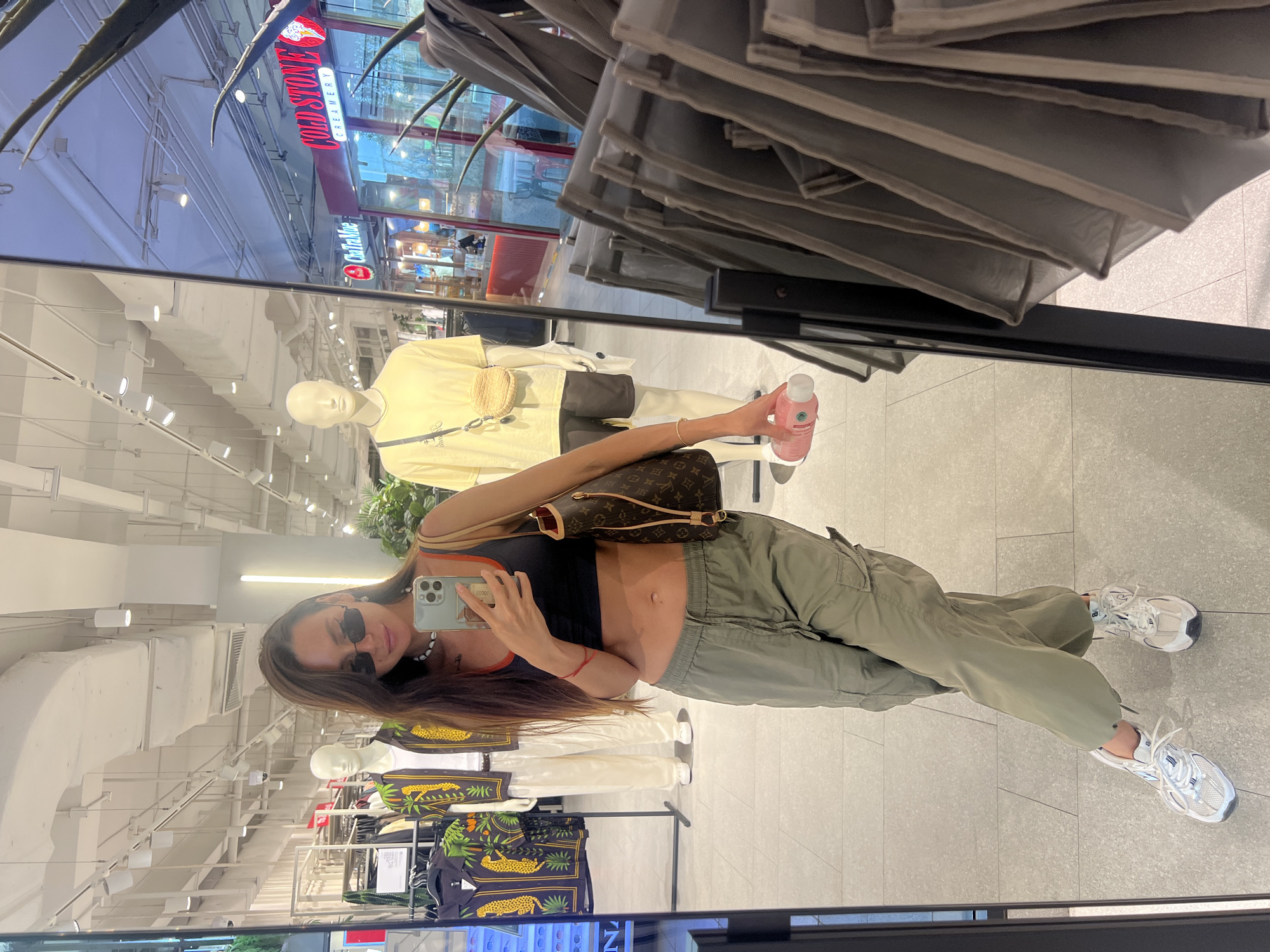 A woman with long brown hair, wearing black sunglasses, a black crop top with orange trim, olive green cargo pants, and white sneakers, stands in a store, taking a mirror selfie. She is holding a pink bottle in her left hand and a brown Louis Vuitton bag over her right shoulder. In the background, mannequins are displayed wearing clothing. Behind the mannequins, store signage for "Cold Stone Creamery" and "ChaTraMue" is visible, suggesting the location is a shopping mall or a retail area. The floor is tiled, and the store features bright overhead lighting.