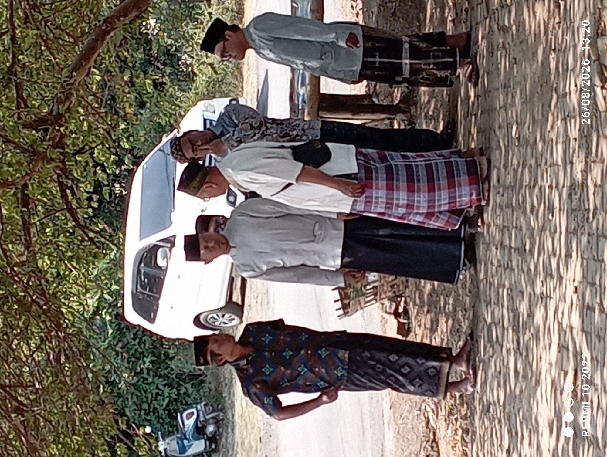 Six individuals, all adult males, are gathered outdoors on a paved surface beneath a canopy of tree branches. The individuals are dressed in traditional attire. From left to right, the first man wears a patterned short-sleeved shirt and a sarong. The second man is in a light-colored long-sleeved shirt and a dark sarong. The third and fourth men are partially obscured, but appear to be in similar attire. The fifth man is dressed in a light-colored long-sleeved shirt and a sarong with a plaid pattern. The sixth man, on the far right, is wearing a light-colored long-sleeved shirt and a dark sarong with vertical stripes.
In the background, a white SUV is parked on a road. To the left of the SUV, a blue and white scooter is visible. There are also several wooden structures, possibly tables or benches, and some foliage. The lighting suggests it is daytime, with shadows cast on the ground from the trees. The image also includes a watermark in the lower left corner that reads "REDMI 10 2022" and in the lower right corner "26/08/2026 13:20".
While the image does not explicitly state the location within Kecamatan Pati, Indonesia, the presence of traditional Islamic attire, the type of vehicles, and the general environmental setting are consistent with many rural or semi-rural areas within Indonesia.