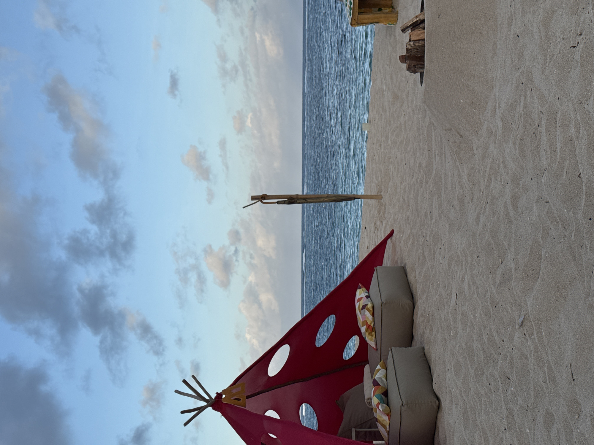 Captured near Kuta Selatan, Indonesia, the scene presents a beachfront view. A red, tent-like structure with circular cutouts dominates the foreground, paired with neutral-colored seating and patterned pillows. Beyond lies a sandy beach leading to a body of water, possibly the ocean, under a sky with scattered clouds.