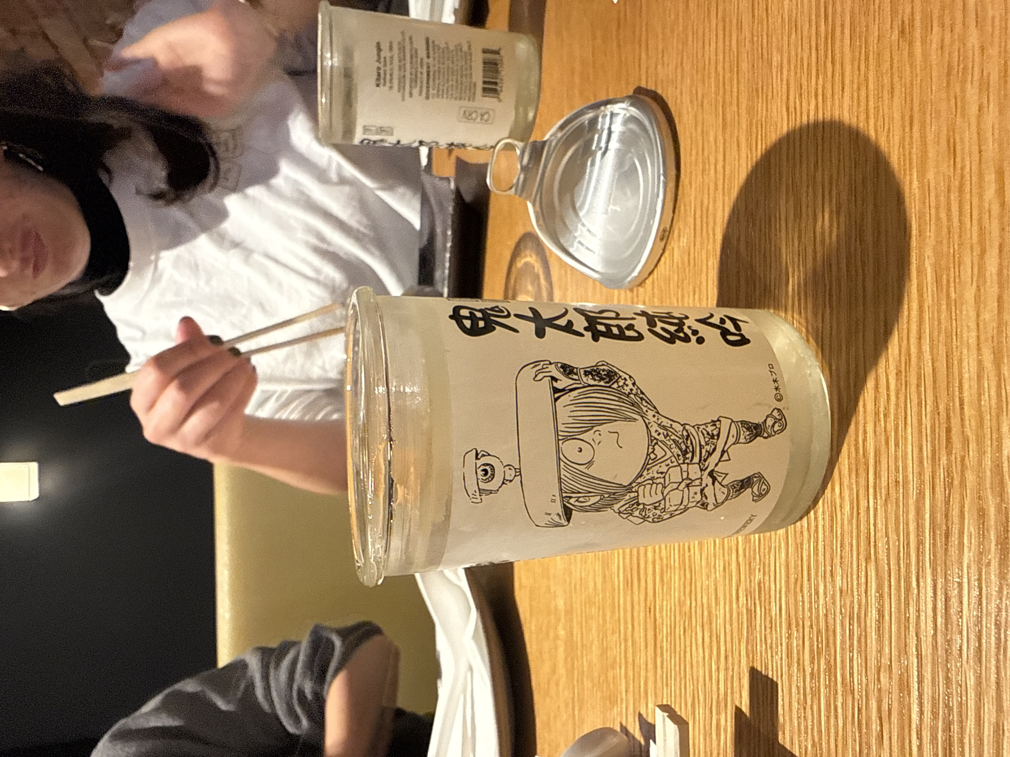 A slightly over-the-shoulder shot shows a person using chopsticks while sitting at a wooden table. Two glass bottles with labels are visible on the table. One bottle appears to be empty, with its lid nearby. The location is suggested to be in New York City, United States. The image's composition focuses on the interaction between the person, the chopsticks, and the bottles on the table, indicating a dining or social setting.