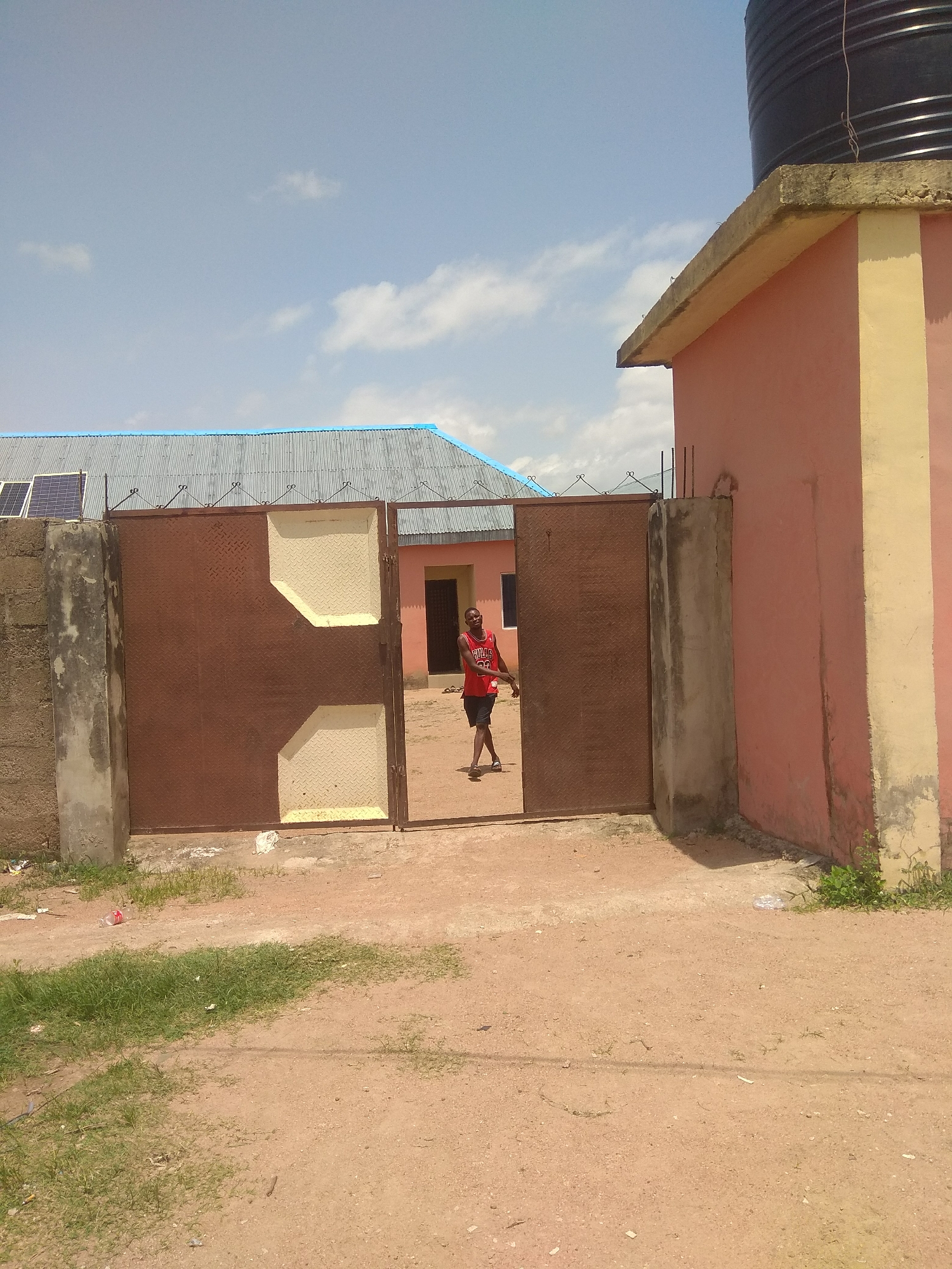 A young man wearing a red Bulls jersey and black shorts walks through a brown metal gate in Jalingo, Nigeria. The gate is partially open, revealing a pink building with a yellow pillar to the right, and a corrugated metal roof with blue trim to the left. Solar panels are visible on the roof. The ground is a dirt path with patches of green grass. A large black water tank is on top of the pink building. The sky is blue with scattered white clouds.