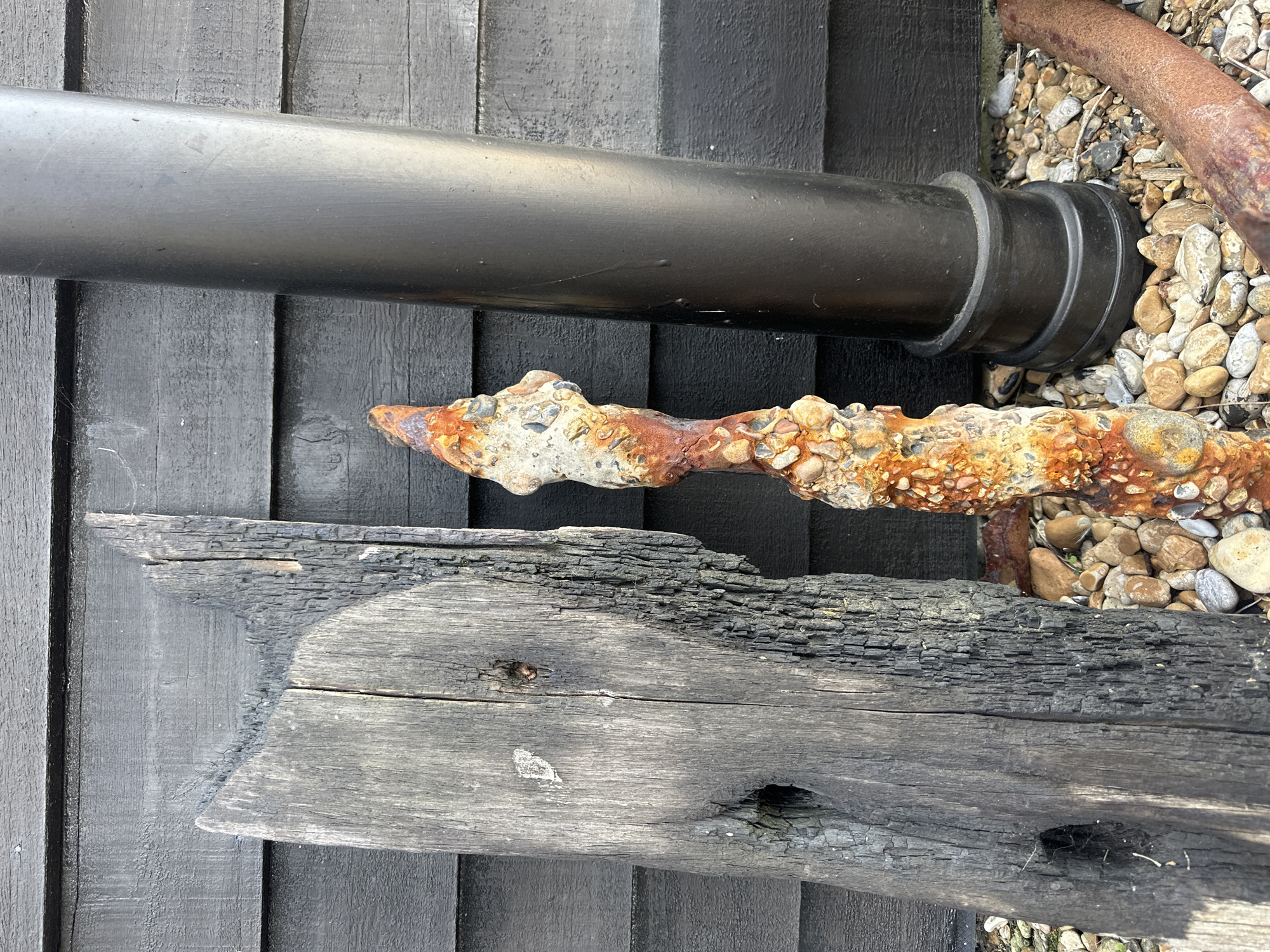 The image shows a close-up view of various objects. A black pipe runs horizontally across the top of the frame, positioned against a dark wooden wall constructed of vertical planks. Below the pipe is a piece of rusted metal, encrusted with small pebbles or shells. Further down is a piece of charred wood, displaying signs of weathering and damage, including holes and cracked surfaces. Small pebbles surround the rusted metal piece.
The location is Folkestone and Hythe District, United Kingdom.