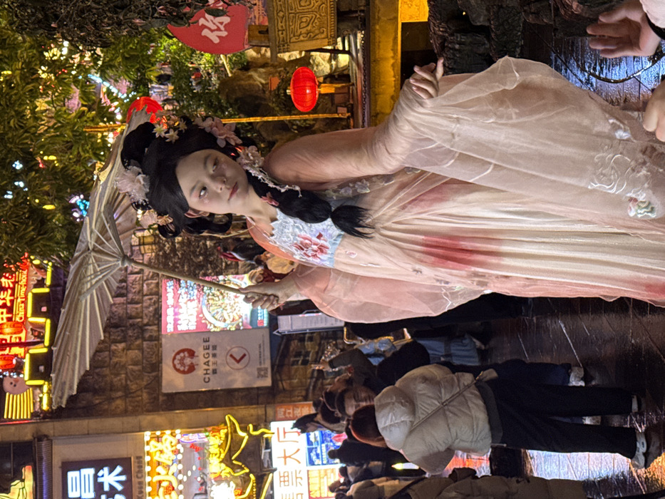 A young individual, likely female, with pale skin and pink cheek makeup, stands center-frame on a wet, reflective dark pavement. They are dressed in an elaborate Hanfu-style costume consisting of a peach-pink sheer outer robe with embroidered floral patterns on the sleeves, a light blue inner bodice with red floral embroidery, and a cream-colored pleated skirt with red accents. Their hair is styled in a black, intricate updo adorned with pink and white floral ornaments and a long braid over their left shoulder. They hold a light-colored, open parasol with a wooden handle in their right hand, extended upwards and to their right. Their left hand is posed with extended fingers near their waist. The individual looks slightly to their right with a serene expression.
Behind the individual, an urban street scene at night is visible. To the left, brightly illuminated commercial signs in Chinese characters read "昌乐艺术" (Chang Le Art) and "售票大厅" (Ticket Hall). A vertical white billboard with a red logo for "CHAGEE 霸王茶姬" (a tea brand) is affixed to a stone-textured wall. Several blurred individuals in contemporary clothing populate the mid-ground, including one person in a white puffer jacket seen from behind. To the right, a decorative landscape features artificial rockwork, illuminated foliage, and red lanterns. A yellow-lit structure is partially visible in the upper right background. The wet ground reflects the ambient lights. This scene is located in a bustling commercial and cultural area within the Yuzhong District, China, consistent with the appearance of Hongyadong.