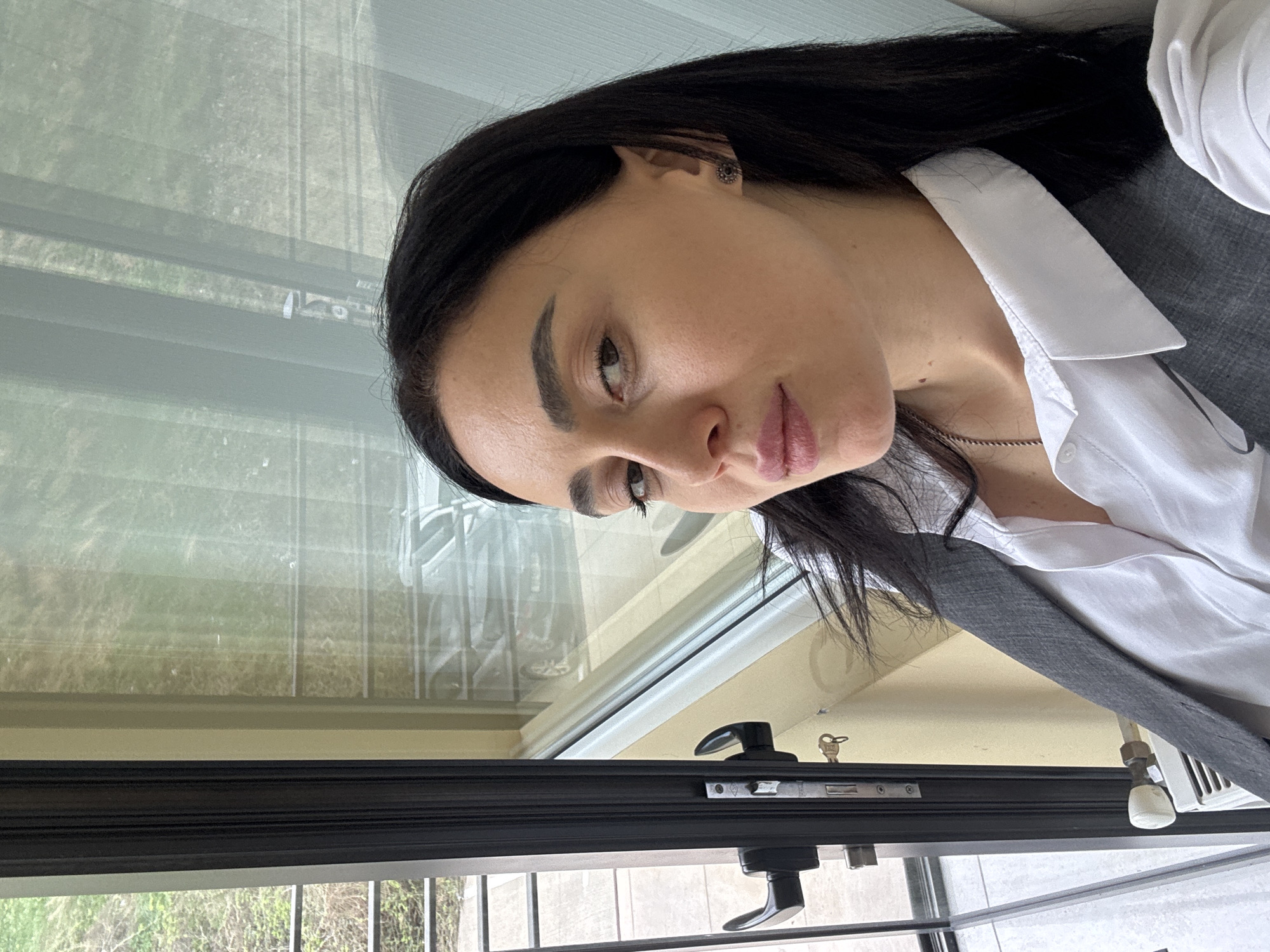 A woman is standing next to a window in Tbilisi, Georgia. She is wearing a white shirt and a grey vest. The window has a black frame and a handle. Outside the window, there are trees. The woman has dark hair and dark eyebrows. She is looking at the camera. She is wearing an earring.