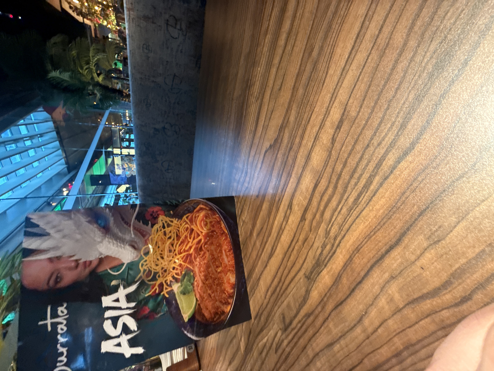 The image shows a table in what appears to be a restaurant in Sofia, Bulgaria. A menu for a restaurant called "Durrata Asia" is on the table. In the background, through a window, there are palm trees and a view of what appears to be a modern building with many windows, suggesting a city environment. The table is made of wood.