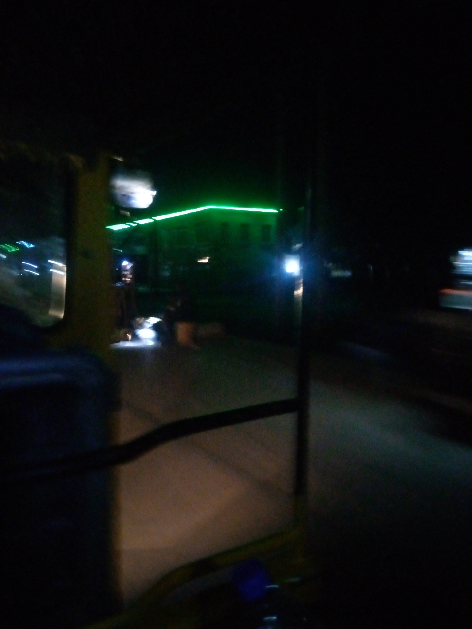 The image depicts a nighttime scene, possibly from inside a vehicle, showing a street in Jalingo, Nigeria. A building with bright green neon lights illuminating its upper facade is visible in the background. The lights cast a glow on the surrounding area, and some streaks of light suggest movement or motion blur. The foreground shows the interior of a vehicle, with a dark dashboard and window, hinting at the perspective of a driver or passenger. The overall ambiance is dark, with artificial lights creating contrasting bright areas.