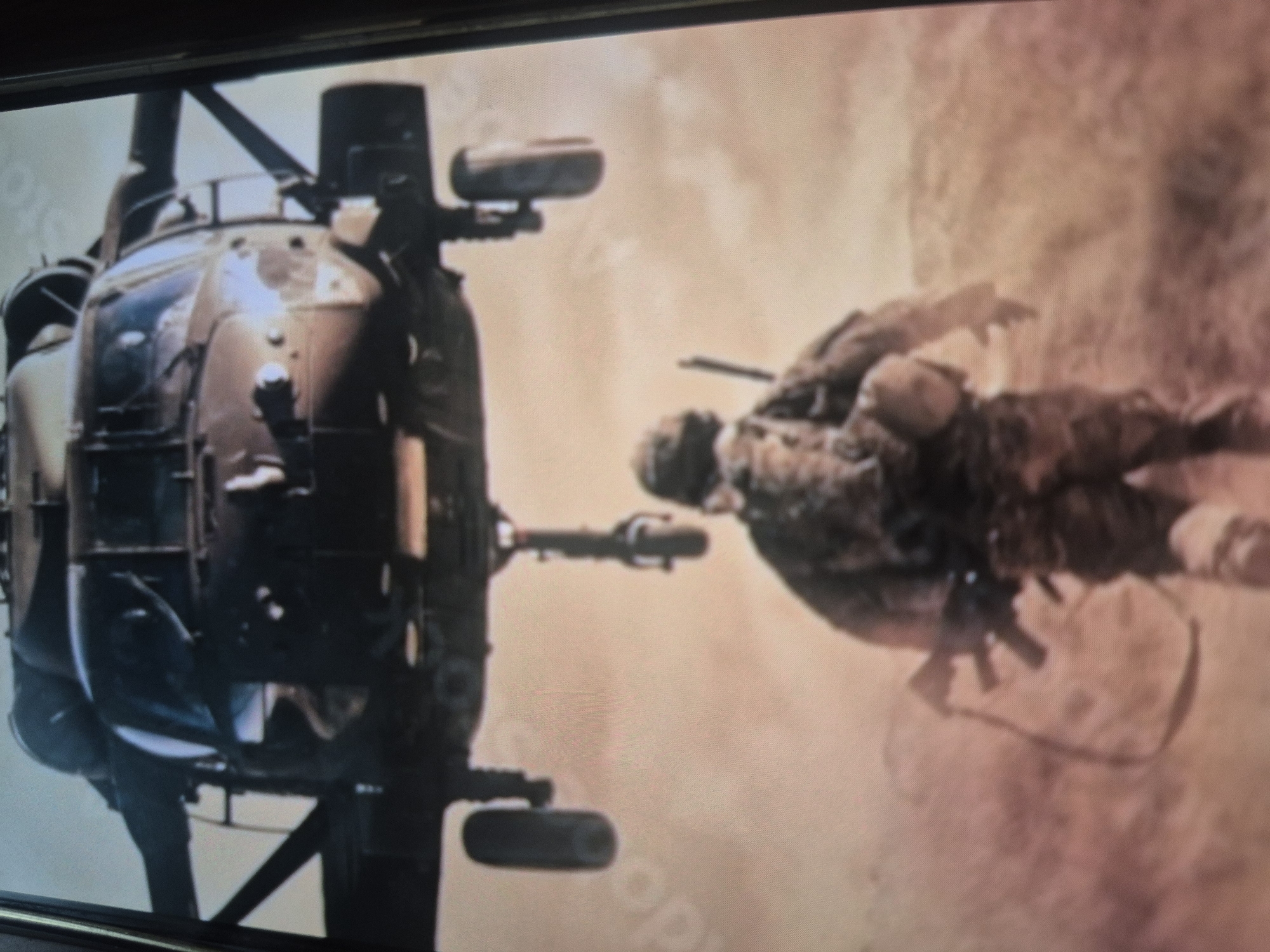 A medium shot captures a soldier falling out of a helicopter, positioned against a dusty background. The helicopter is on the left, angled to show its side. The soldier is on the right, positioned horizontally as if falling. He wears military garb and carries a rifle. The background appears to be an arid, sandy environment. The location in Tbilisi, Georgia is not evident from the visual information provided.