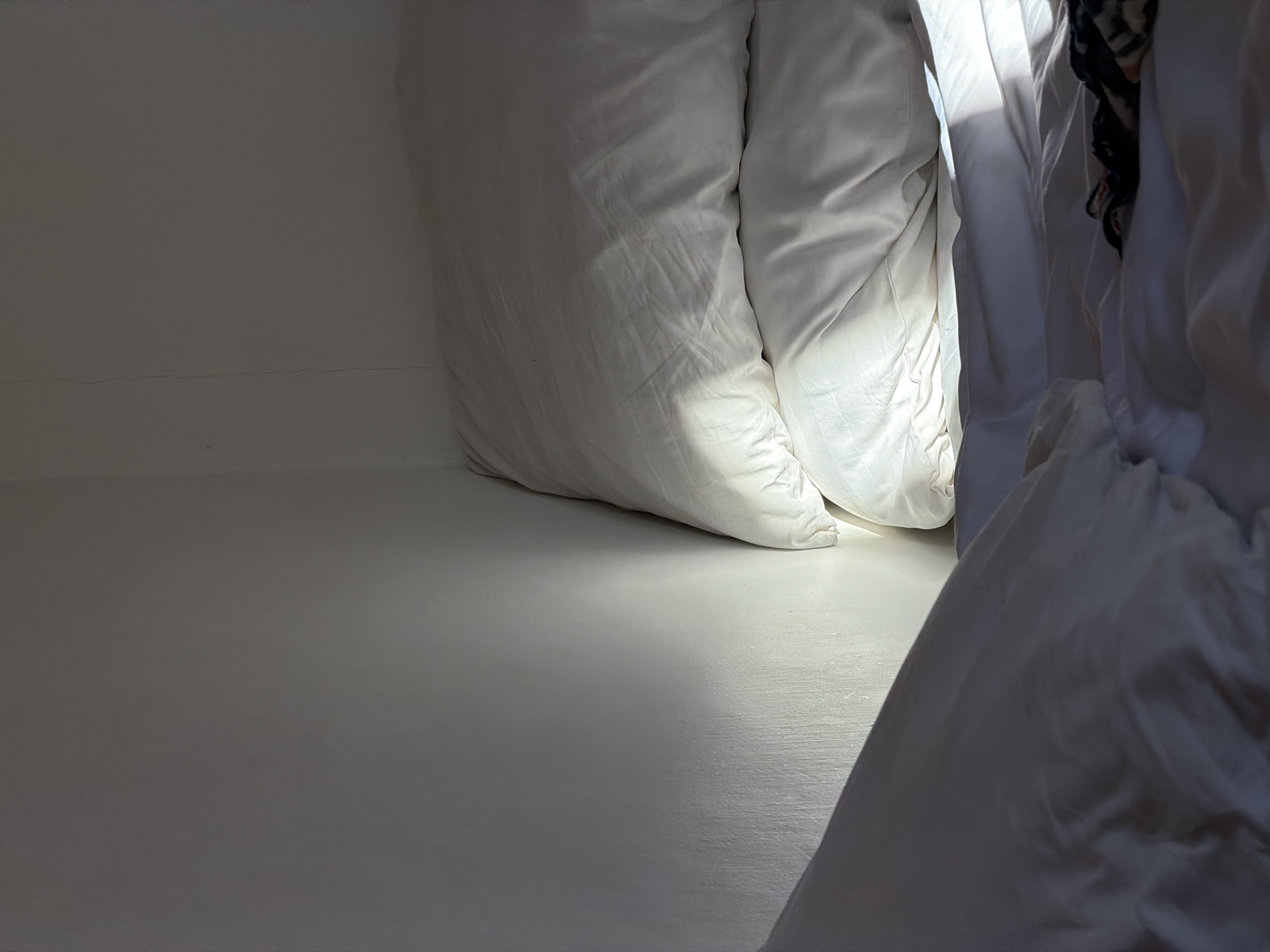 The image shows a low-angle view inside a small, likely enclosed, space, possibly in North Kuta, Indonesia, though this cannot be verified visually. The primary elements are white bedding, including pillows and draped fabric, positioned against a white wall. The space is brightly lit, with a beam of light illuminating the floor and the bedding. No people are visible. The focus is on the play of light and shadow on the white surfaces. The wall appears to be lightly textured.