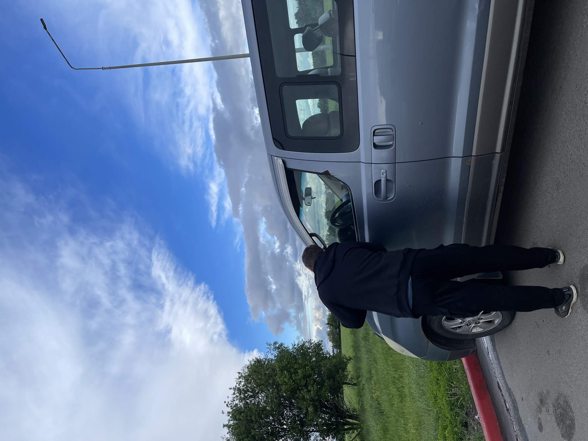 A man in a dark-colored hoodie and pants is standing next to a silver van, leaning forward and looking into the driver's side window. The van is parked on the side of a road next to a grassy area with trees. The sky is blue with scattered clouds. A tall light pole is visible in the background. The location is near Бавыкино, Russia.