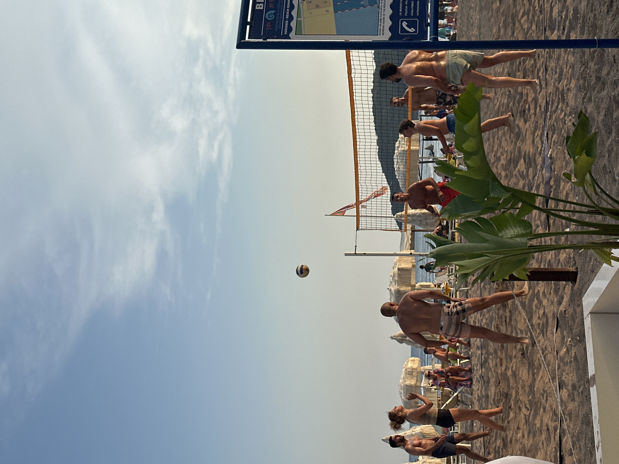 This image depicts a beach volleyball game on a sandy beach in Boreti, Montenegro. Several individuals are actively playing volleyball, with some positioned on either side of a net. A volleyball is visible in mid-air. In the background, beach umbrellas and lounge chairs are set up, suggesting a recreational area. The sea and a tree-covered hill are visible in the distance. A blue signpost with text and a map is present on the right side of the frame. The sky is clear with some scattered clouds. The overall scene is of a lively beach activity on a sunny day.