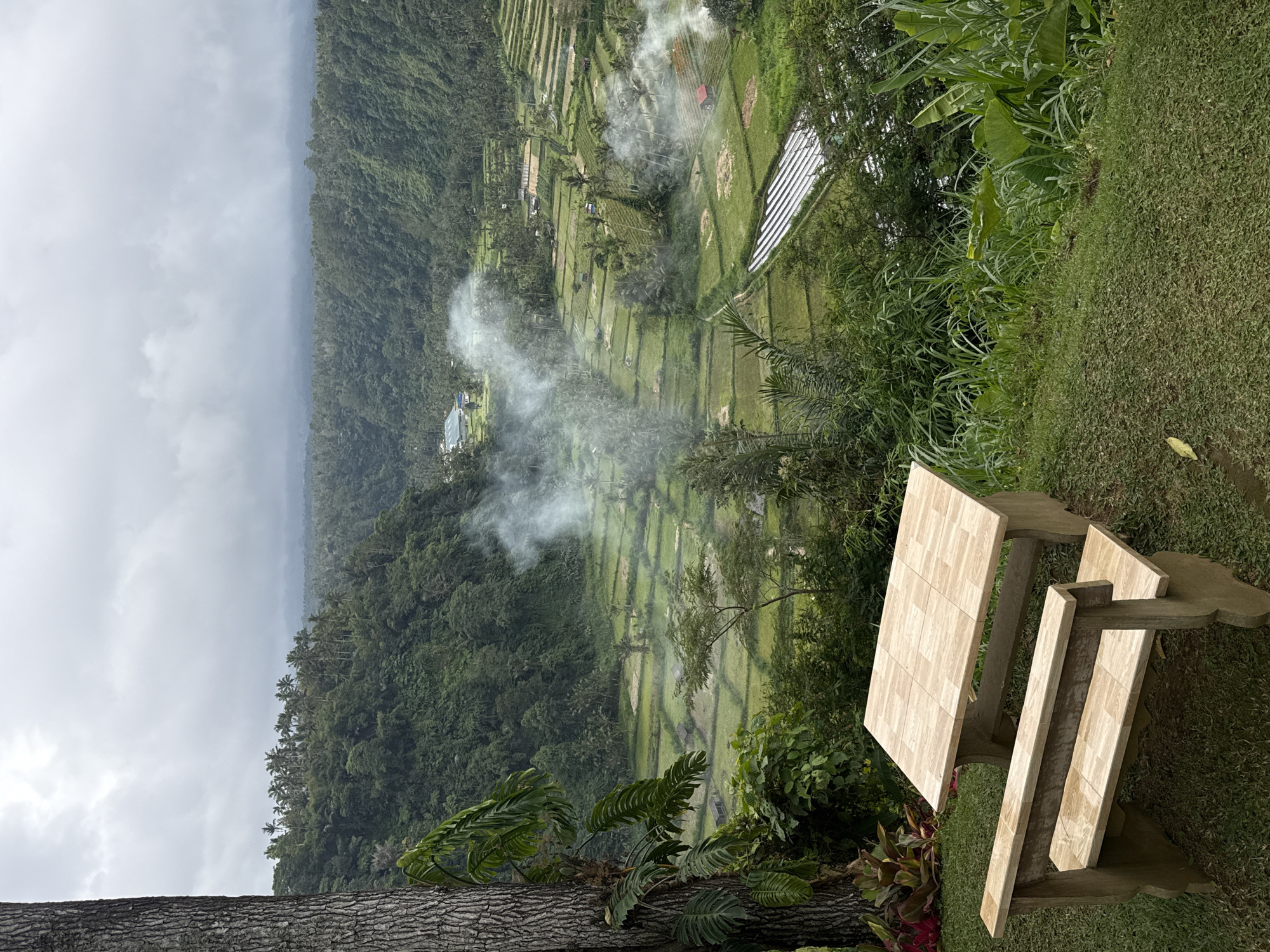The image depicts a panoramic view of lush green rice terraces cascading down a valley in Rendang, Indonesia, under a cloudy sky. In the foreground, a wooden picnic table and bench sit on a grassy knoll, offering a vantage point for the scenery. Smoke drifts through the valley from various points, possibly indicating agricultural activity or cooking fires. The terraces are interspersed with palm trees and small structures, suggesting a rural settlement. The overall impression is one of natural beauty and tranquil rural life.