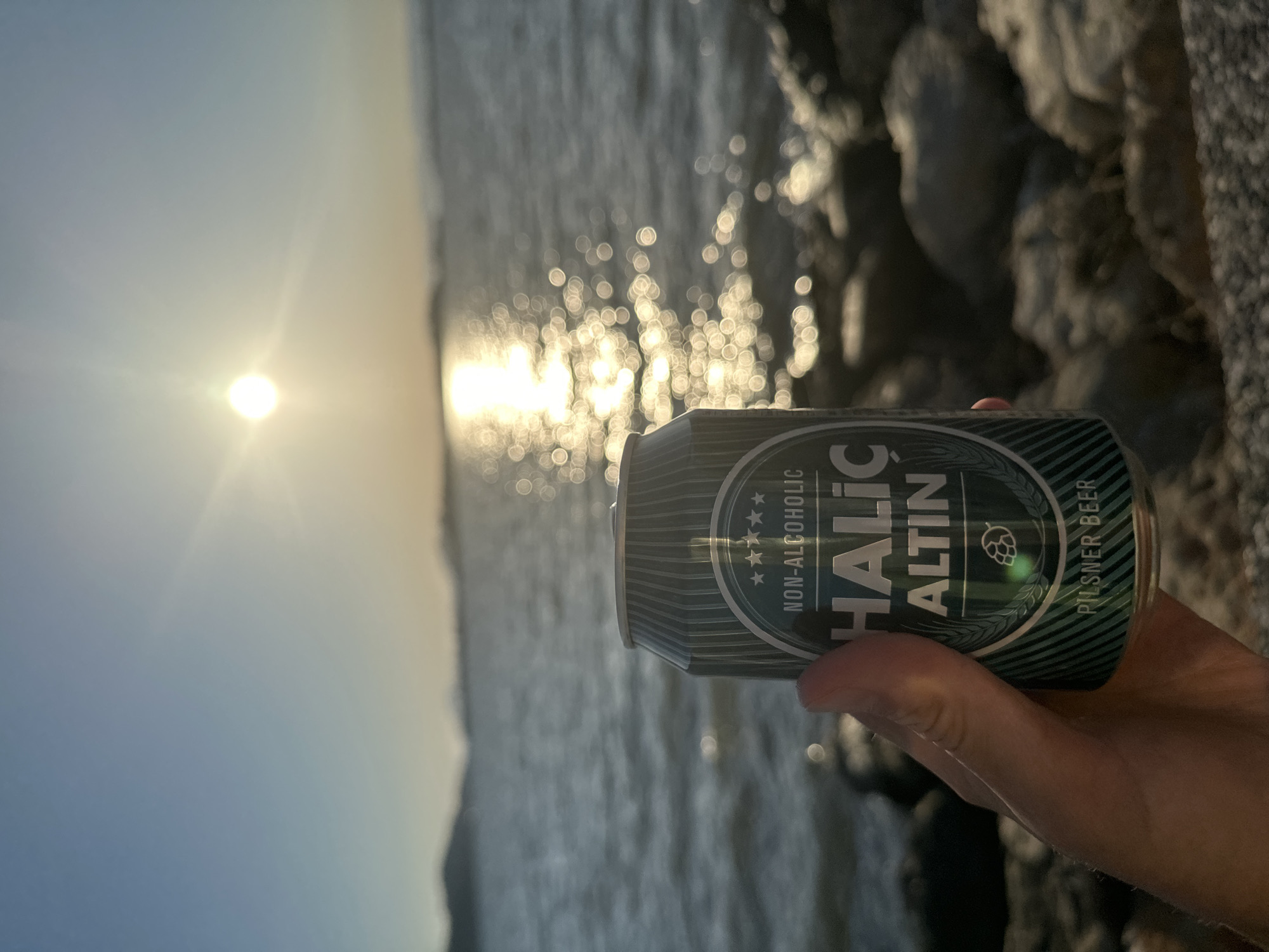 A person's hand holds a gray and green non-alcoholic beer can against a scenic backdrop of water at sunset in Fethiye, Turkey. The sun, a bright orb, is low on the left side of the frame, casting a shimmering, golden reflection path across the water. The water appears slightly choppy, creating numerous bright, circular bokeh effects from the sun's reflection.
On the right side of the image, a dark, textured rocky shoreline or seawall is visible, partially blurred. The sky above the horizon is a clear, light blue, transitioning to a warmer, brighter hue near the sun. The overall lighting suggests either late afternoon or early evening.
The can is held by a hand, with fingers wrapped around it, showing skin and some arm hair. Visible text on the can includes "NON-ALCOHOLIC", "HALİÇ ALTIN", and "PİLSNER BEER", along with a stylized hop cone logo and five stars. The design of the can features diagonal stripes in various shades of green and gray. The image captures a serene moment of enjoying a beverage by the water during a beautiful sunset.