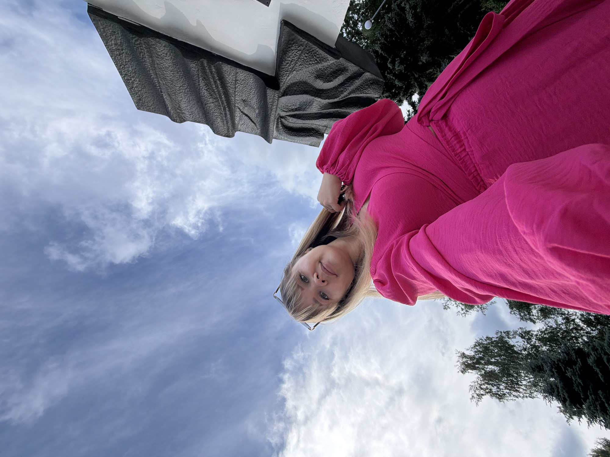 A low-angle, upward shot shows a young woman in a vibrant pink dress and glasses, looking up at the camera. She is positioned in the lower right quadrant of the frame, with her right hand touching her hair. Behind her, a dark, textured monument rises against a sky filled with wispy white clouds. To the left of the monument, a tall, slender tree and a dark green evergreen tree are visible. The woman's pose and the upward perspective suggest she is standing near a monument in Chaikovsky, Russia.
