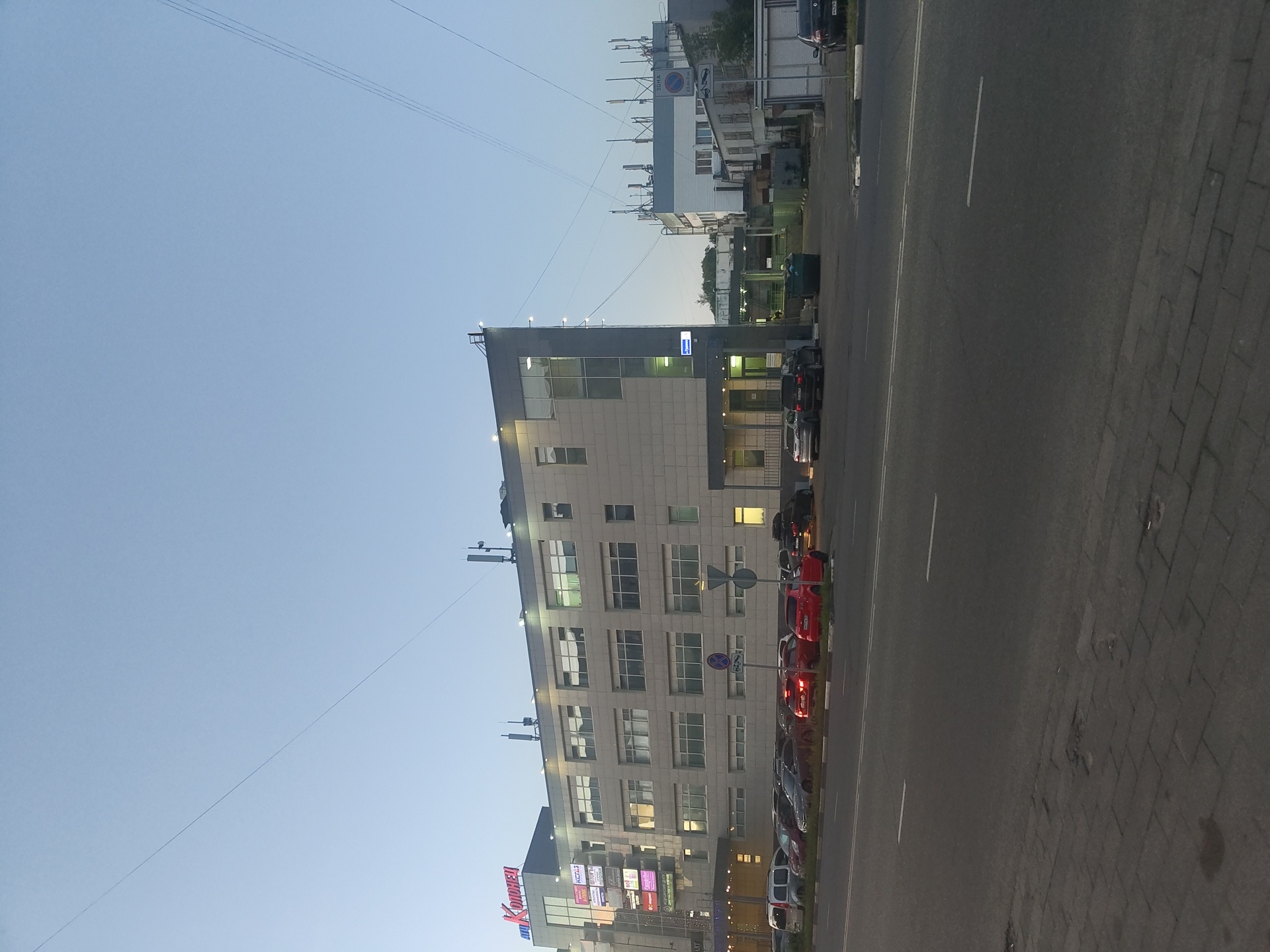 The image shows a modern, multi-story commercial building with a light-colored facade, situated on a street in Zhukovsky, Russia. The building has numerous large windows and appears to be a shopping center or office complex, as indicated by signage that includes the name "АТЦ КОЛОНЕЦ" in Cyrillic letters at the top. In front of the building, a paved parking lot is filled with various cars, including sedans, SUVs, and a white van. The street in the foreground has a paved sidewalk and asphalt roadway with lane markings. Electrical wires are visible strung across the sky, and several telecommunication antennas are mounted on the rooftops of the buildings. The sky is clear and pale blue, suggesting it is either early morning or late evening. There are no people visible in the image.
