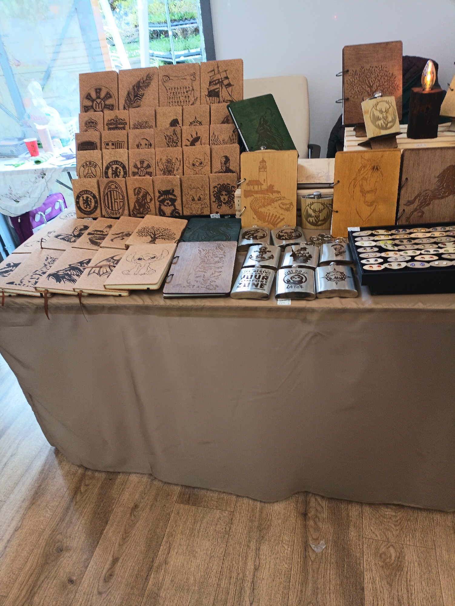 The image shows a table at what appears to be a market stall. The table is covered with a tan cloth, and displays a variety of handcrafted items including notebooks with various designs engraved on their covers, hip flasks with engraved designs, and a tray of small, round objects, possibly pins or earrings. The designs on the notebooks and flasks feature a mix of symbols, animals, logos, and nature scenes. In the background, there is a window with a view of greenery, and part of a building is visible. The setting may be a fair or craft market in Burgas, Bulgaria. The flooring suggests an indoor location, potentially a community center or indoor market hall.