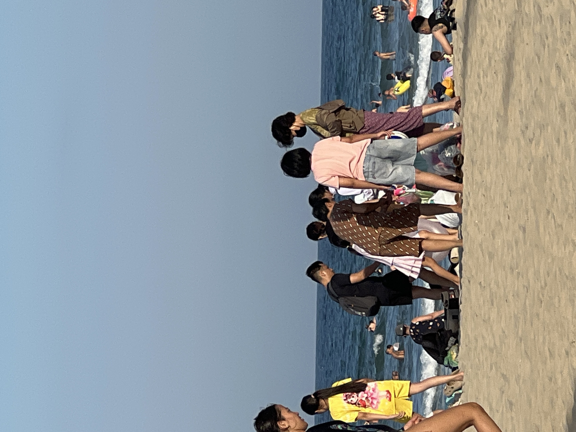 The image depicts a busy beach scene in Đà Nẵng, Vietnam. Several groups of people are present, enjoying a sunny day by the ocean. The foreground is dominated by a sandy beach, with a rough texture indicating the dry sand.
In the midground, a line of people are either sitting or standing, facing the water. They are dressed in casual summer clothing, including t-shirts, shorts, and skirts. Some individuals are wearing hats. There are also various items of clothing and bags scattered on the sand near them.
The background features the blue ocean with gentle waves. Several people are visible in the water, some appearing to be swimming or wading. A yellow inflatable float is also present in the water. The sky is a clear, light blue, suggesting fair weather and likely a daytime scene, possibly late morning or afternoon.
The overall atmosphere seems relaxed and recreational, with people enjoying a typical beach day. The presence of numerous individuals and scattered belongings suggests a popular spot. No text is visible in the image.