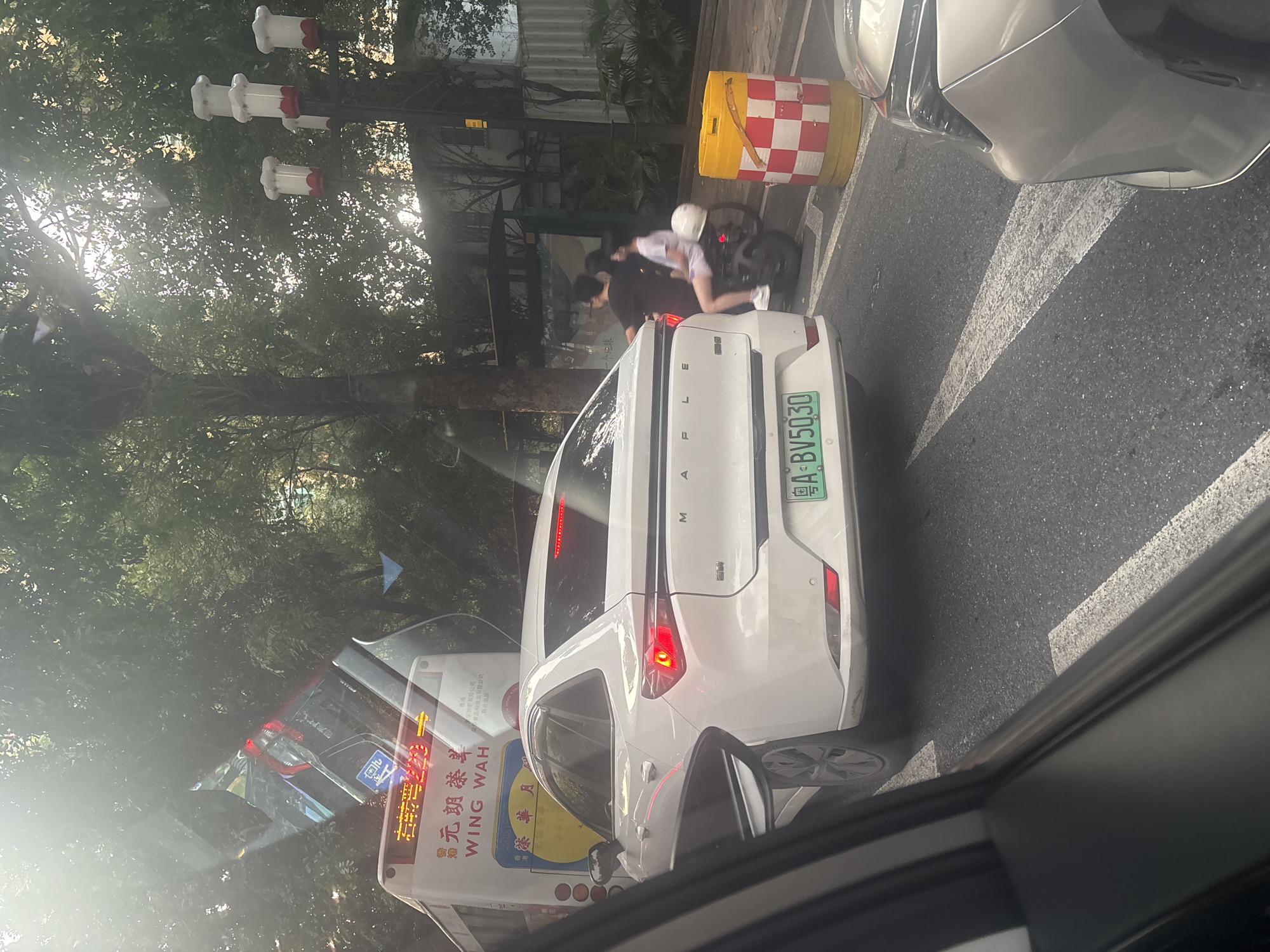 The image captures a street scene in Haizhu District, China. A white sedan, identified by the license plate "A·BV5030", is parked on the right side of the road, facing away from the camera. The car's brake lights are illuminated. To the left of the sedan, a bus with Chinese characters and the text "WING WAH" is visible, suggesting it might be a public transport vehicle. In the background, several people are seen, including a couple on a motorcycle. The environment features lush green trees, a lamppost with decorative elements resembling chef hats, and streetlights. The scene appears to be a busy urban street with traffic.