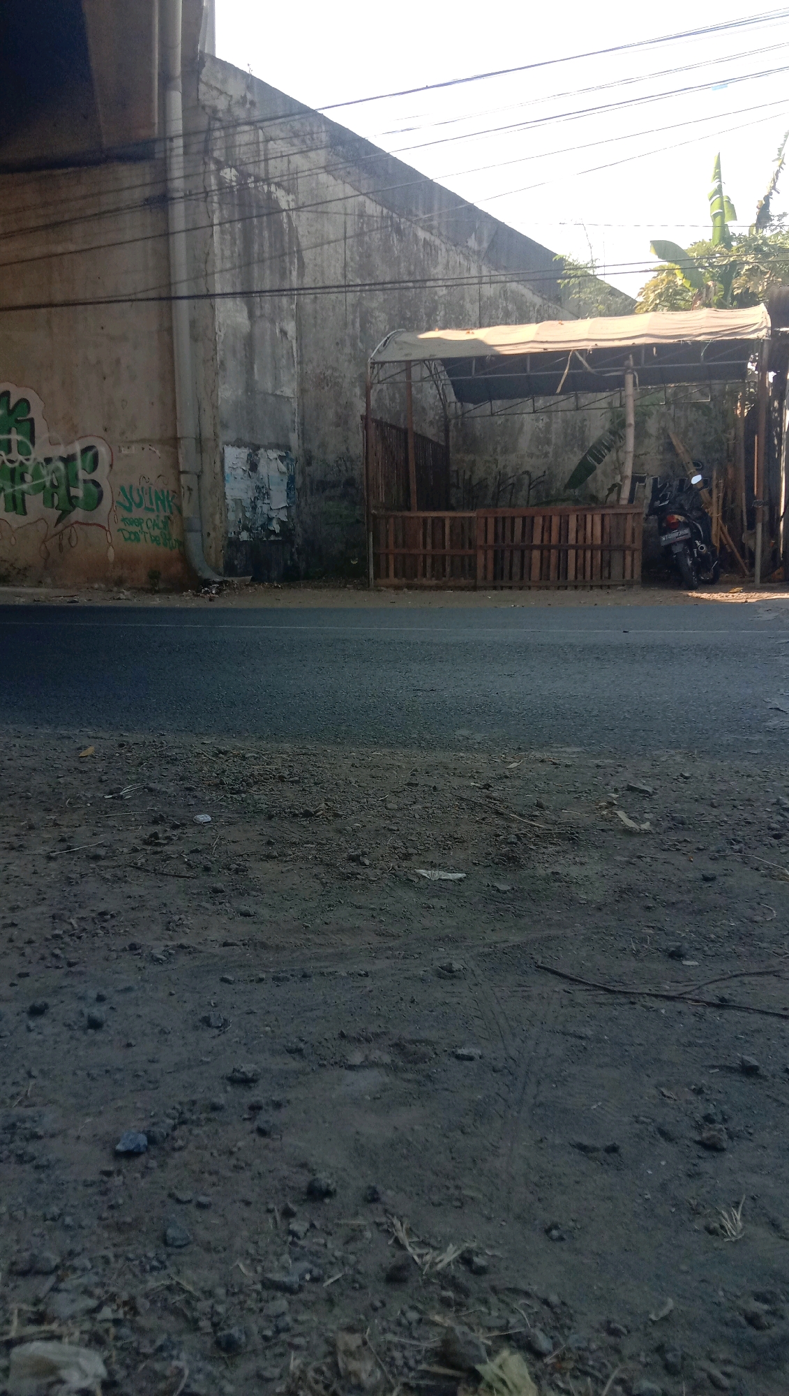 The image depicts a roadside scene under an elevated structure, likely a bridge or overpass, in Karanganyar, Indonesia. The elevated structure, made of concrete and metal, casts a shadow over a significant portion of the scene. Beneath the structure, a weathered concrete wall runs horizontally. Graffiti, predominantly in green spray paint, is visible on the lower section of the wall.
Adjacent to the wall, a makeshift shelter with a light-colored tarp roof is erected. In front of this shelter, a wooden fence, constructed from what appears to be pallet wood, forms a barrier. Behind the fence and within the shelter, vegetation, including what looks like banana leaves, is present. A black motorcycle is parked to the right of the shelter, partially obscured by the wooden fence.
The foreground of the image is dominated by a paved road, which appears to be asphalt. The surface of the road shows some wear and tear, with scattered debris like small stones and dry vegetation. The ground immediately off the road consists of dirt and gravel, also with visible debris. Power lines are strung across the top of the image, running parallel to the elevated structure. The lighting suggests it is daytime, with sunlight illuminating the upper portions of the scene.