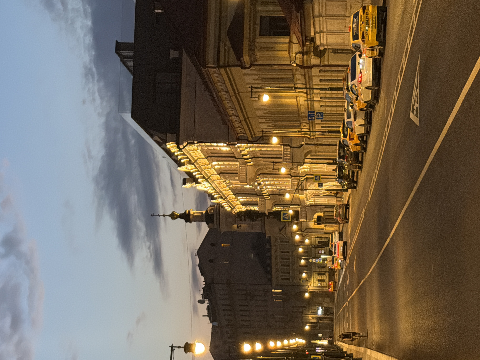 The image captures a twilight scene on a city street, likely in Moscow, Russia, with a wide asphalt road in the foreground leading towards buildings illuminated by warm, yellow streetlights. The sky is a dusky blue with scattered clouds. On the right side of the street, a line of parked yellow taxis are visible. On the left side of the street, a lone cyclist is pedaling away from the camera. The buildings lining the street feature ornate architecture and are lit from within and by exterior lighting, suggesting it is evening. The atmosphere is calm and quiet, with no visible interactions between any of the people or objects.
