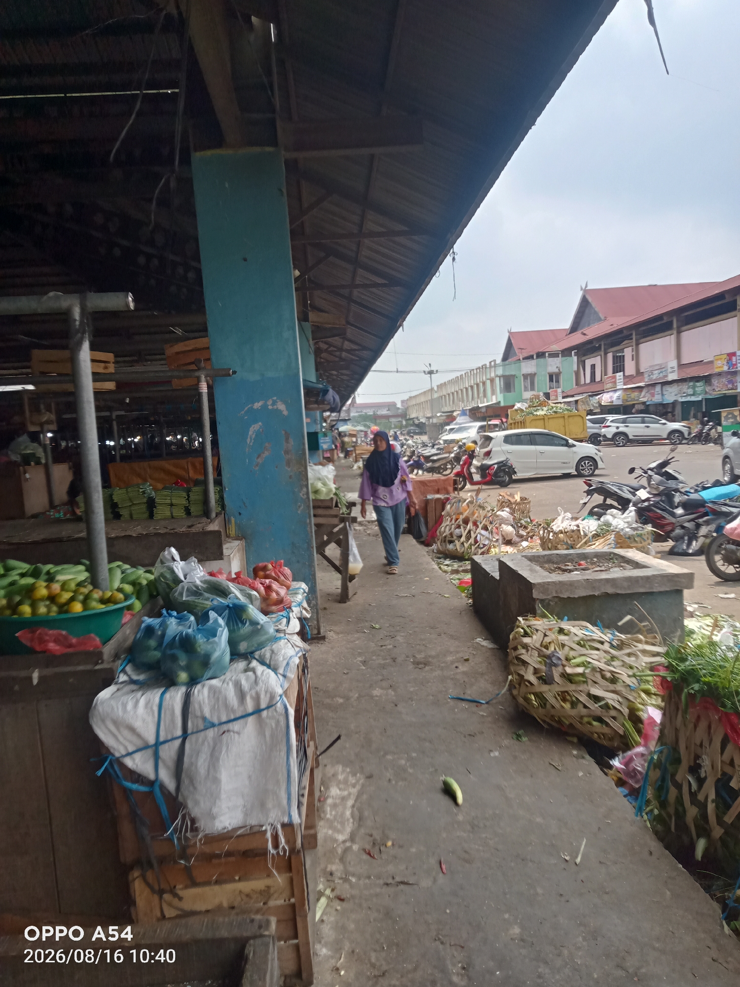 The image captures a street scene in Jambi, Indonesia, likely a market area. A woman wearing a hijab and a purple shirt is walking away from the camera, down a paved pathway. To her left, there are market stalls with various produce, including cucumbers and what appear to be oranges in a green basin, as well as produce bagged in blue plastic. Behind the stalls, there are more bags of produce and some items wrapped in white cloth. To the right of the path, there are large woven baskets filled with green produce, possibly vegetables, and concrete structures. Further down the street, a variety of vehicles, including cars and motorcycles, are parked or moving. Buildings with red roofs are visible in the background. The environment appears to be an open-air market with covered sections. The overall lighting suggests it is daytime.