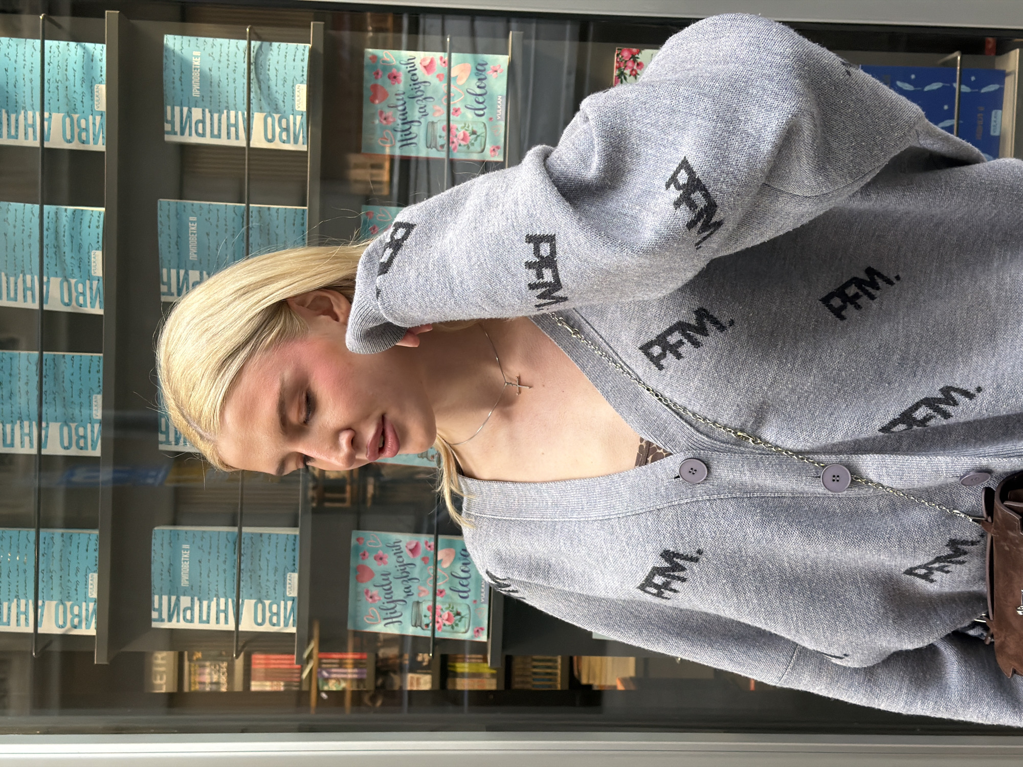 A blonde woman, with her head tilted downwards and eyes closed or cast low, is depicted in what appears to be a contemplative moment. She is wearing a grey knitted cardigan adorned with repeated black "PFM." lettering and fastened with light purple buttons. A delicate silver chain with a small cross pendant hangs around her neck, and a brown bag with a silver chain strap is visible on her right side.
She stands in front of a glass display case filled with books. The books are primarily in Serbian, indicated by the Cyrillic script. Multiple copies of works by "ИВО АНДРИЋ" (Ivo Andrić), a prominent Serbian author, are visible, specifically "ПРИПОВЕТКЕ I" and "ПРИПОВЕТКЕ II" (Short Stories I and II). Another prominent title is "Hiljadu razbijenih delova" (A Thousand Splendid Suns). The publisher "VULKAN" is also clearly visible on several book spines and covers. The scene suggests an indoor setting, likely a bookstore or a display window in Belgrade, Serbia, consistent with the language of the books. The lighting appears to be natural daylight.