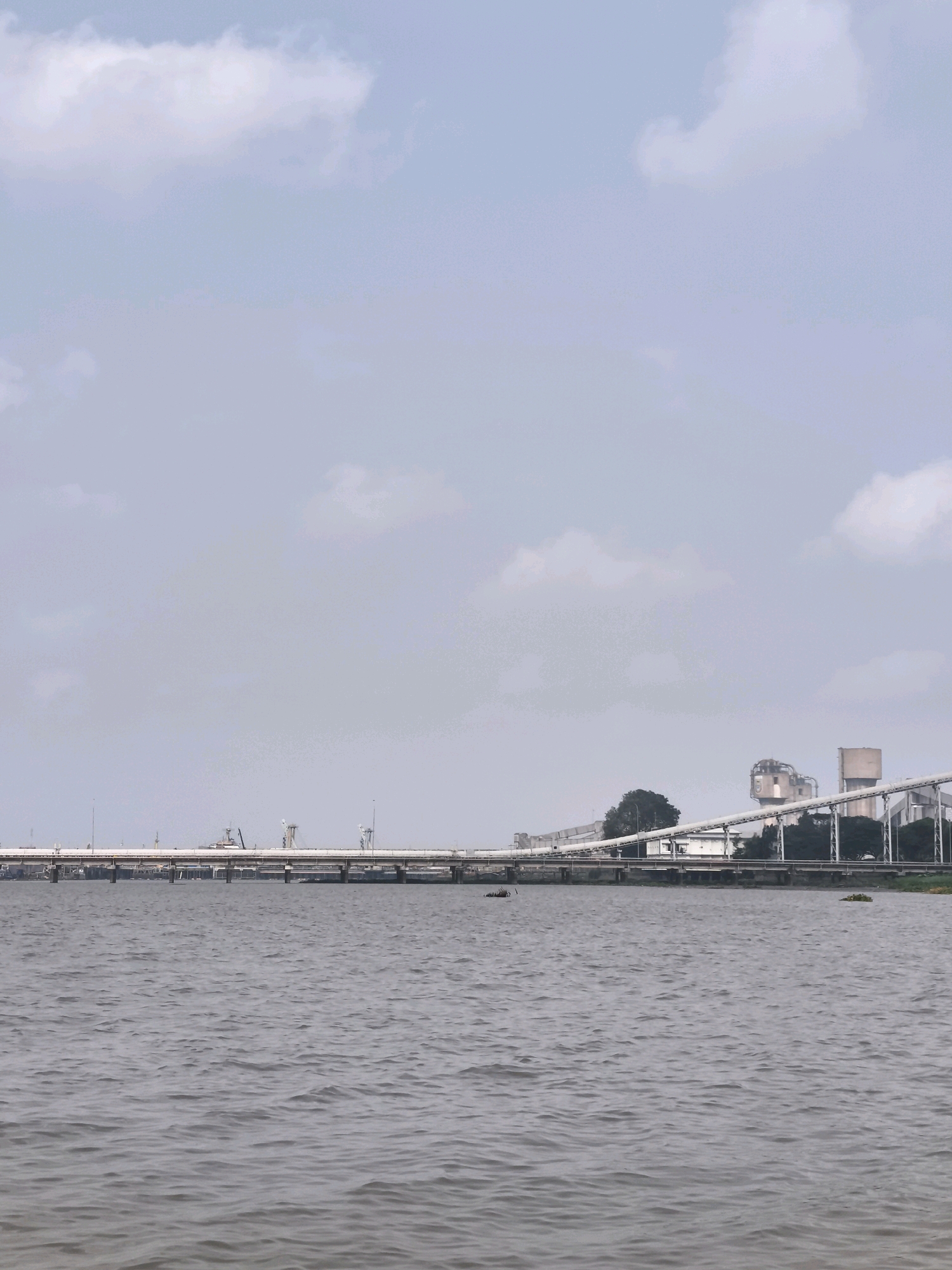 The image depicts a wide, calm body of water in the foreground, likely a river, with gentle ripples on its surface. In the mid-ground, a long bridge stretches horizontally across the water, supported by numerous pillars. The bridge appears to be part of an industrial area. On the far bank, beyond the bridge, a complex of industrial buildings and structures is visible, characterized by silos, conveyor belts, and water towers. These structures suggest an industrial or port facility. Lush green trees and vegetation are interspersed among the industrial buildings. The sky above is partly cloudy, with soft white clouds against a pale blue backdrop. The overall scene suggests an industrial waterfront environment in Palembang, Indonesia, possibly near a port or processing plant.