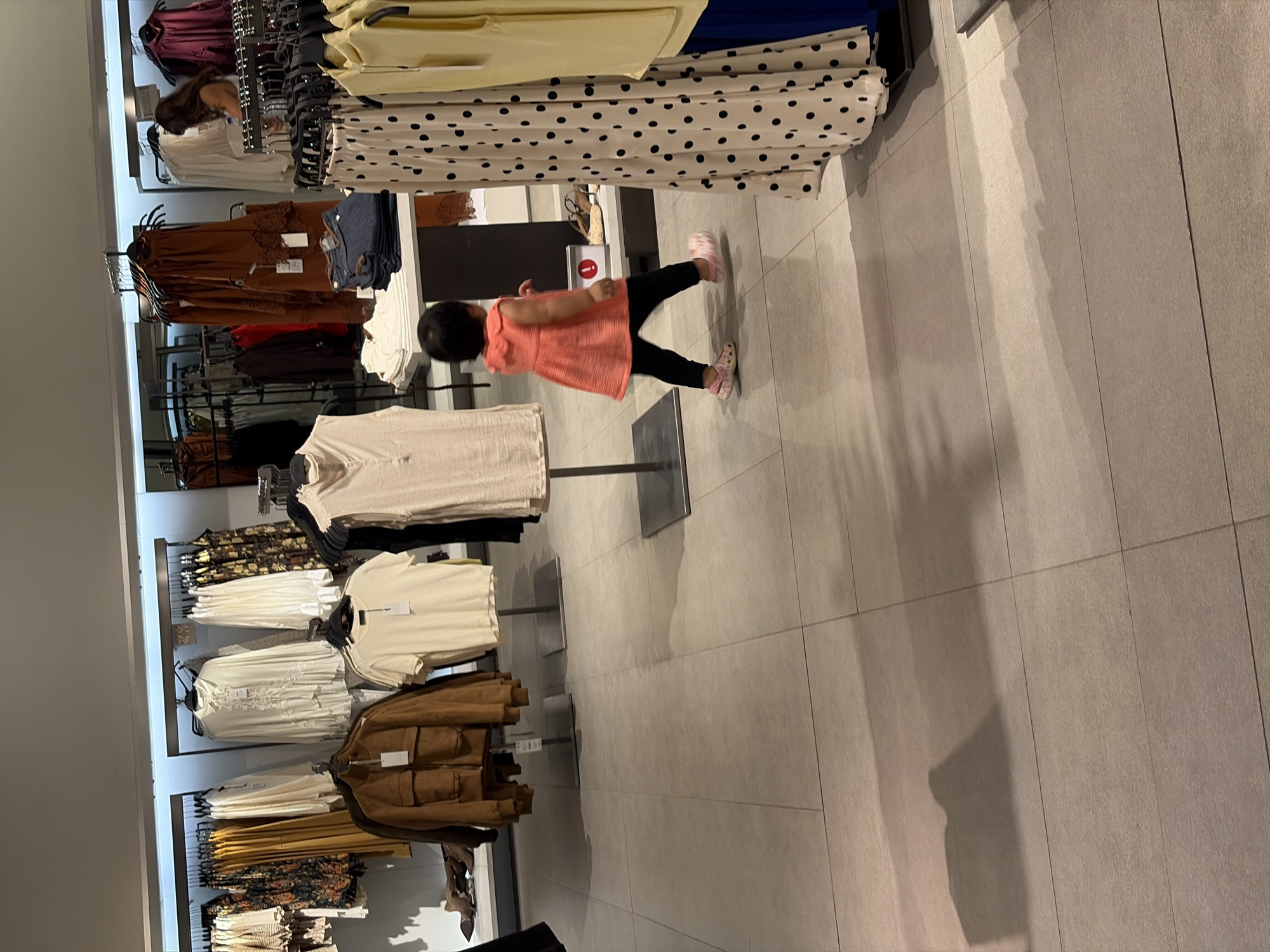 In a retail clothing store in Kuta, Indonesia, a young child in a coral-colored dress and black leggings walks across the tiled floor. The child is facing away from the camera, moving towards the right side of the frame. In the background, clothing racks display various garments, including jackets, dresses, and shirts. A woman is visible further in the background, browsing merchandise. The lighting is artificial, casting shadows on the floor.