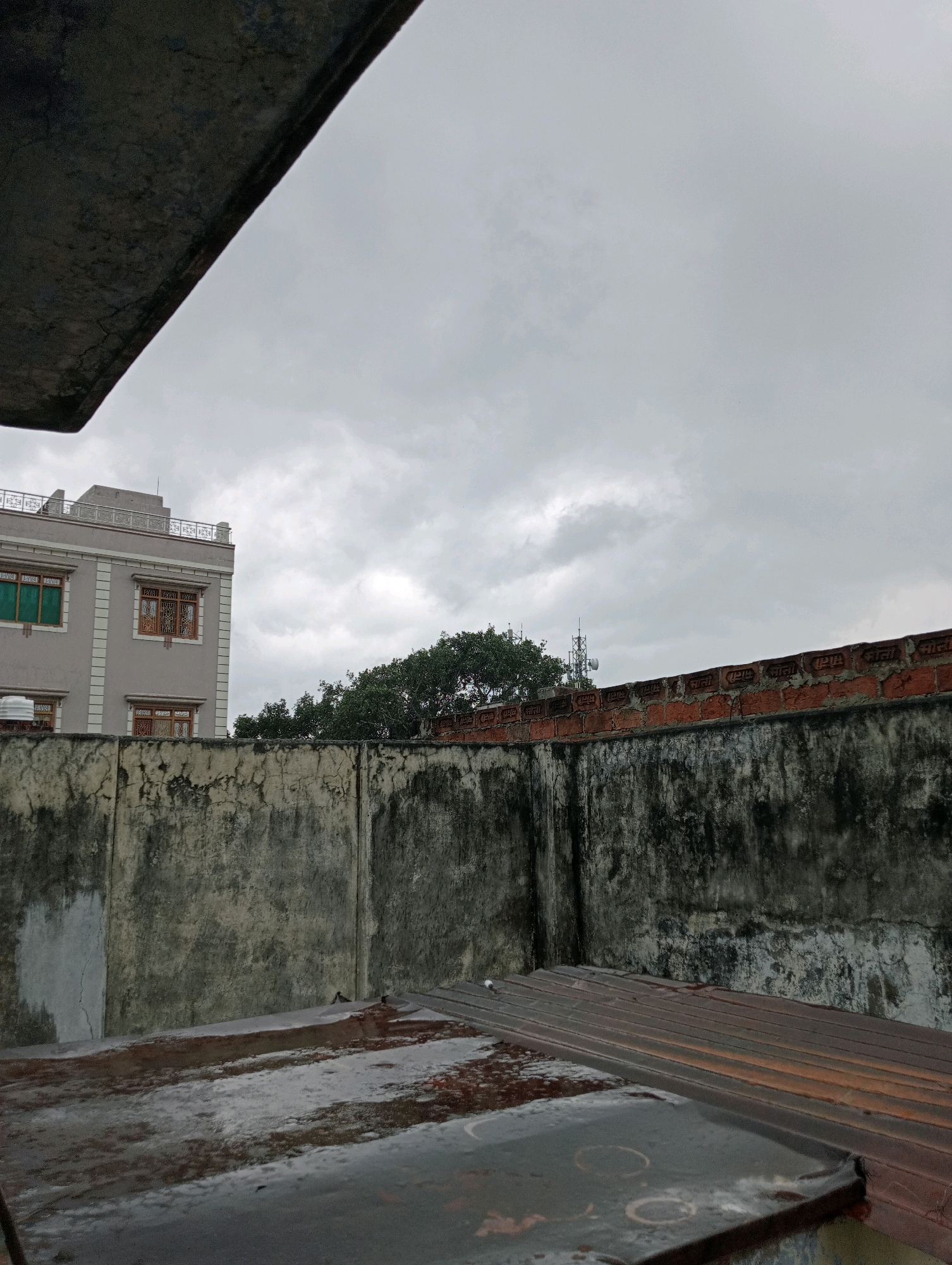 The image depicts a cloudy sky over a city, likely Varanasi, India, as suggested by the architectural style and the presence of a telecommunications tower. In the foreground, there is a rusty corrugated metal roof, partially covered with what appears to be patchy frost or grime. Behind this roof is a weathered, textured concrete wall, with dark stains and cracks, suggesting age and exposure to the elements. Above the concrete wall, a section of a building is visible, characterized by a light-colored facade, multiple windows with wooden frames, and a decorative railing on its rooftop. Further in the background, dense green trees can be seen, and beyond them, a telecommunications tower stands against the overcast sky. The overall scene conveys a sense of urban decay and an approaching or recent rain. There are no people visible in the image.