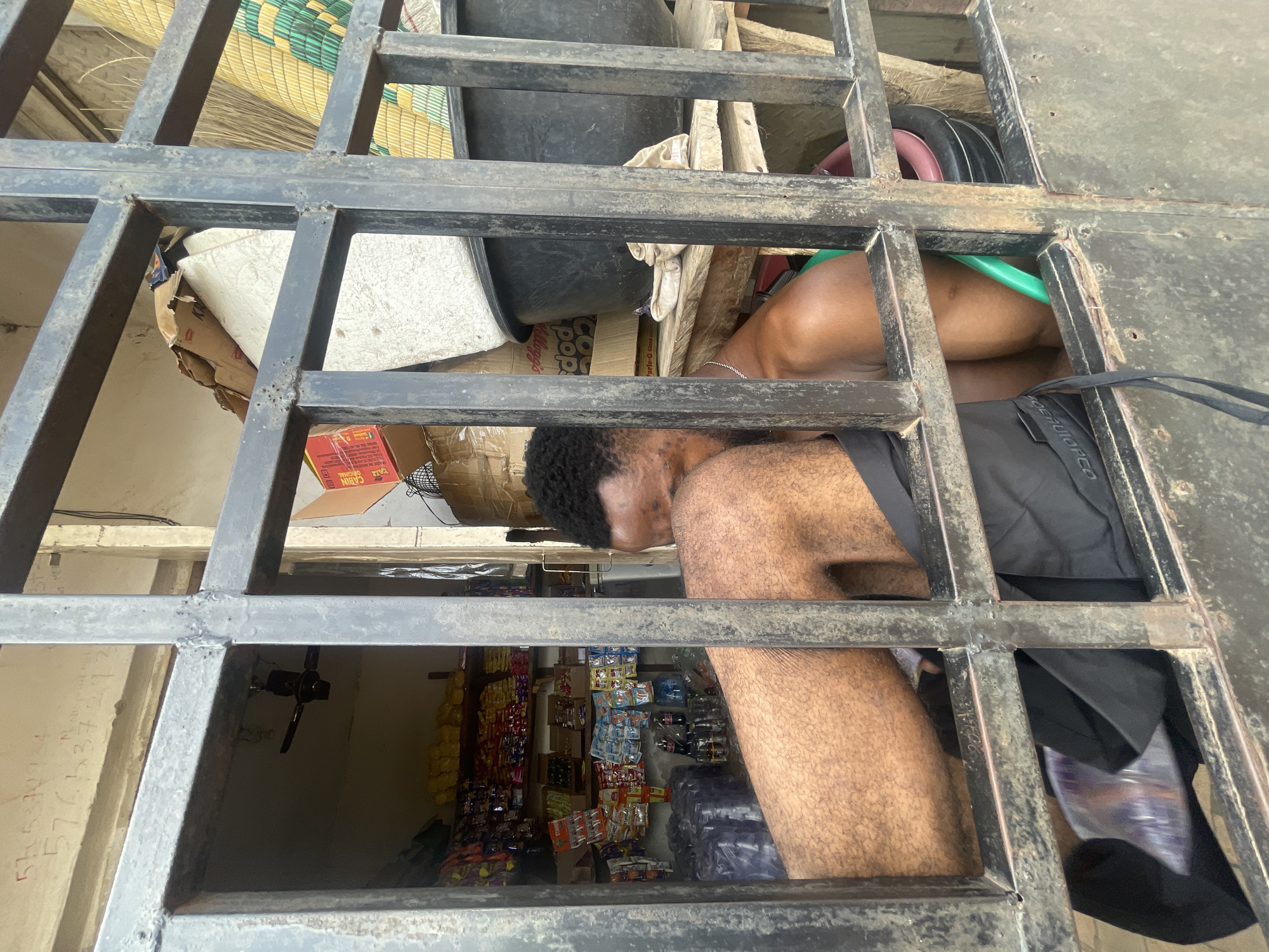 The image shows a man looking through a metal grate, likely a security barrier. He has dark hair and tan skin and is wearing a dark-colored shirt or jacket. The background reveals what appears to be a small store stocked with snacks and drinks, indicative of a commercial environment. It's difficult to pinpoint the exact location within Lokoja without additional landmarks or context. The scene is an everyday view into a typical Nigerian shop.