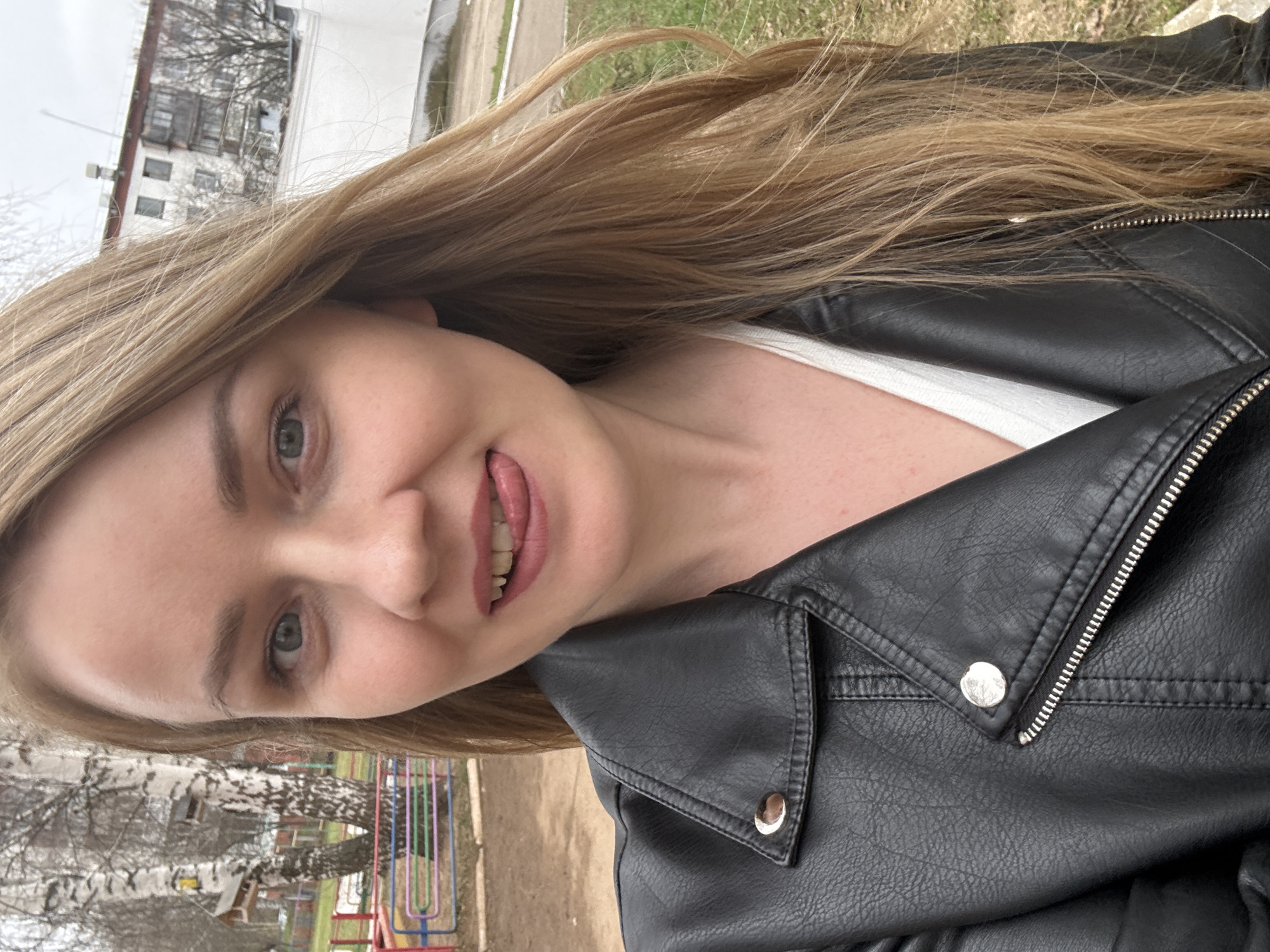 A young woman with long, straight blonde hair and light-colored eyes poses for a selfie outdoors. She is smiling playfully with her tongue slightly extended. She wears a black leather-style jacket with silver zippers and snaps, layered over a white top. The image is taken at a close-up, high-angle, resulting in a tilted perspective.
The background reveals an outdoor setting, likely a park or residential area in Chaikovsky, Russia, during late winter or early spring. Bare-branched trees, including distinctive birch trees with white bark, are prominent. A light-colored, multi-story building, possibly an apartment block, stands in the distance. Colorful playground equipment, resembling climbing bars, is partially visible in the lower right background. The ground appears patchy with dry grass or earth. The weather seems to be overcast, with soft, diffused daylight. No visible text is present in the image.