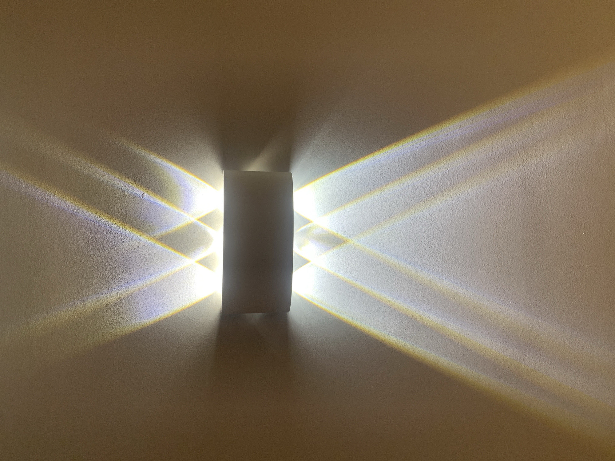 The image shows a wall-mounted light fixture that emits beams of light in a symmetrical pattern, creating an abstract visual effect. The fixture itself is a simple, rectangular prism, positioned vertically in the center of the frame. The light it casts forms distinct, sharp-edged rays that spread outwards and upwards, intersecting to create a diamond-like pattern on the textured, beige-colored wall. Some of the light beams exhibit subtle color gradients, hinting at prismatic effects or the way light interacts with the fixture's material. The wall's rough texture is accentuated by the play of light and shadow, adding depth and dimension to the scene. There is no indication of people, specific weather conditions, or a precise time of day beyond the presence of artificial lighting. The setting appears to be indoors, likely a room with a plain, neutral-colored wall. The overall impression is one of geometric abstraction and the interplay of light, creating an atmospheric and modern aesthetic.