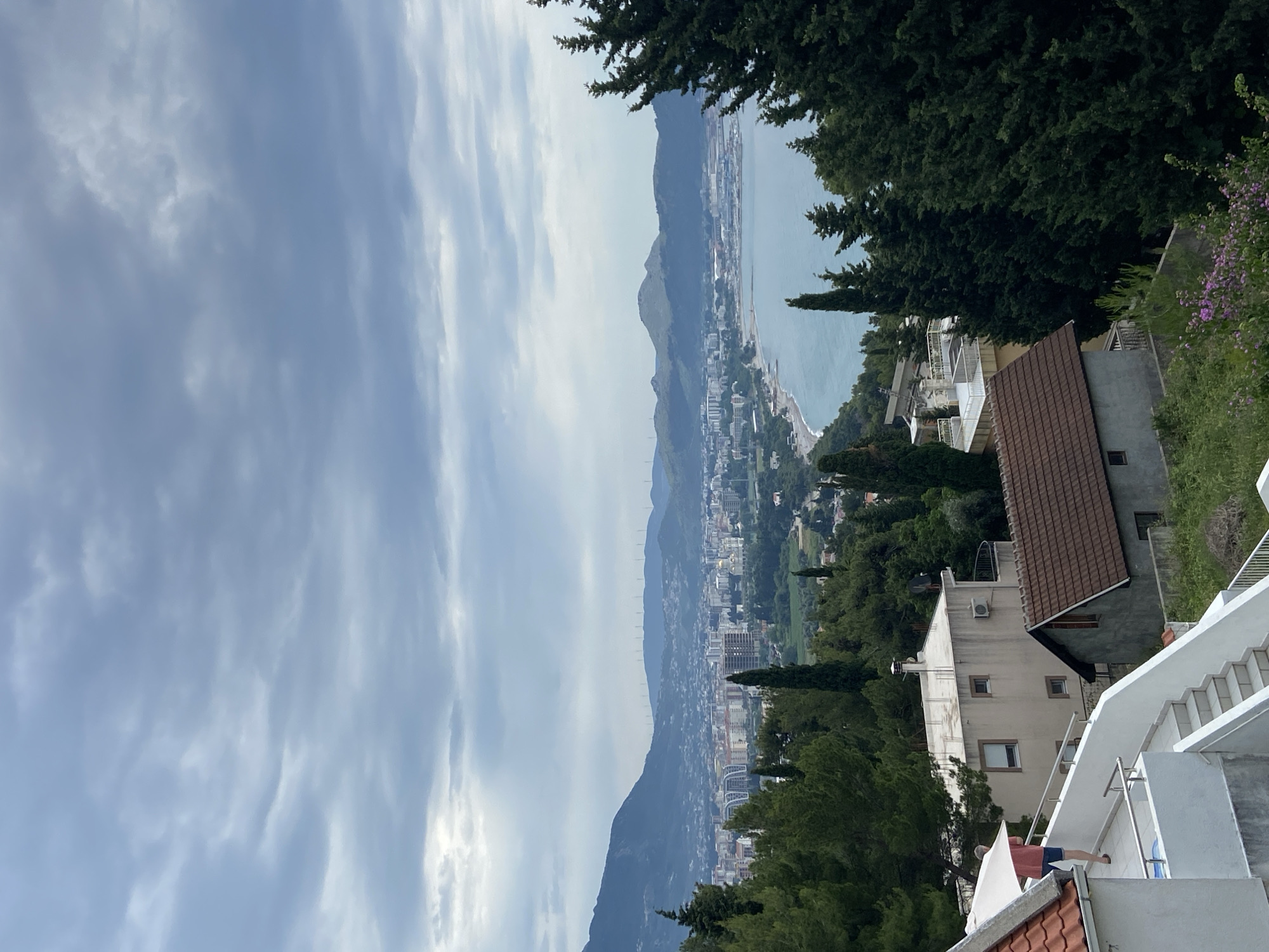 The image is a scenic view overlooking the coastal city of Šušanj, Montenegro. The foreground features residential buildings with red-tiled roofs and white exteriors, including visible staircases and balconies. Verdant trees interspersed among the buildings partially obscure the cityscape.
In the midground, the urban area of Šušanj stretches along the coastline, framed by a backdrop of mountains. The landscape features a mix of residential, commercial, and infrastructural elements.
The background is dominated by a wide expanse of the Adriatic Sea under a cloudy sky.