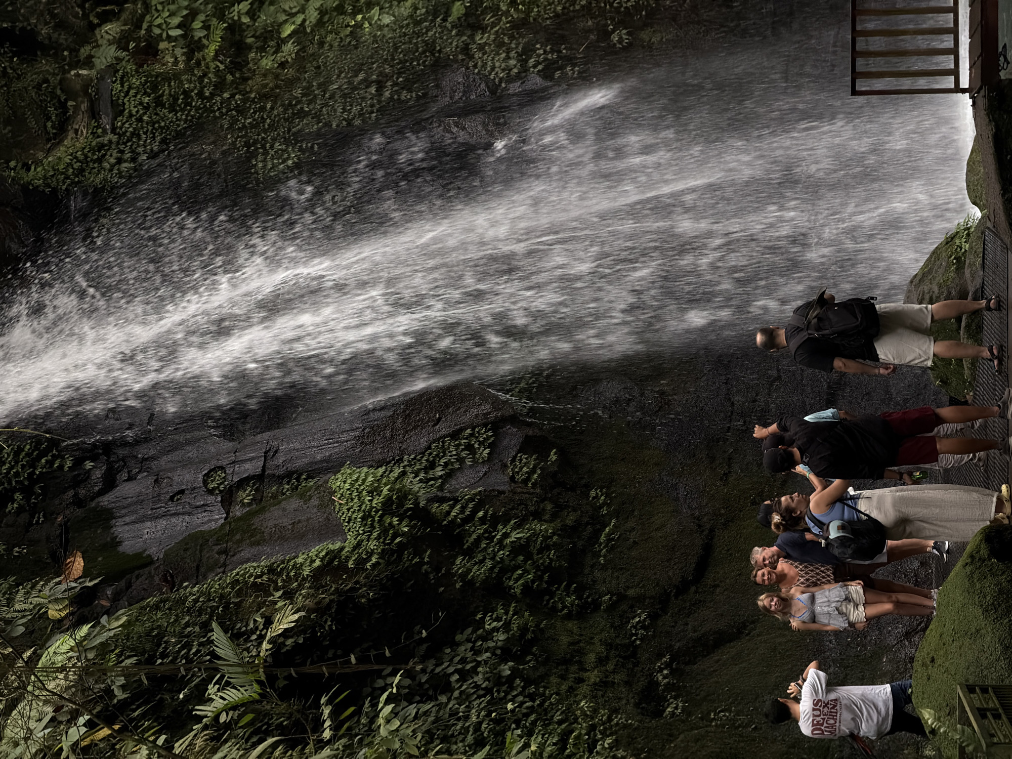 In Tegallalang, Indonesia, a group of people stand near a cascading waterfall. A man in a white t-shirt with the words "DEUS EX MACHINA" printed on it is taking a photo of two women and two men standing in front of the waterfall. The women are dressed in casual clothing, one in a blue and white striped top and white shorts, and the other in a light blue top and white pants. The men are also dressed casually, one in a dark blue shirt and shorts, and the other in a black t-shirt and red shorts. Another person, a woman with a backpack, is standing to the right of the group, looking towards the waterfall. The waterfall itself is a prominent feature, with water flowing down a dark, moss-covered rock face. The surrounding environment is lush and green, with trees and vegetation visible on either side of the waterfall. There is a wooden platform with a railing in the foreground, suggesting a designated viewing area for visitors. The scene appears to be a tourist destination, with people enjoying the natural beauty of the waterfall.