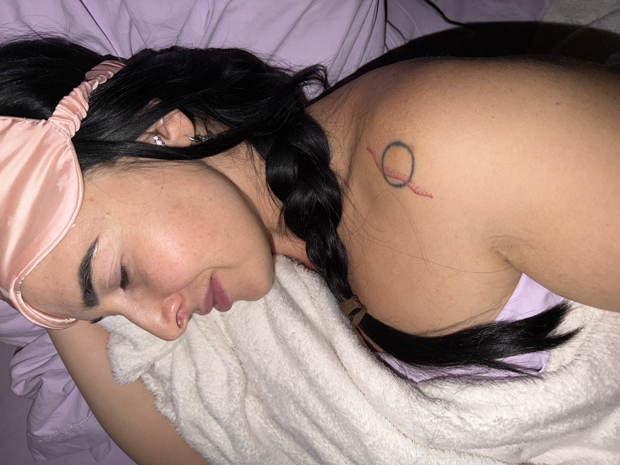 A close-up, high-angle shot captures a person, likely female, resting or sleeping in an indoor setting, possibly a bedroom. Her head and upper body are visible, positioned on a light purple pillow or sheet. She wears a light pink satin sleep mask over her eyes. Her dark, long hair is pulled into a neat braid, secured with a brown elastic band, resting on a fluffy, white blanket or towel.
The person's eyes are closed, and her facial expression is serene, suggesting sleep or deep relaxation. Visible features include a well-defined dark eyebrow, and slightly parted lips. On her ear, multiple piercings are visible: a silver hoop and a cluster of three small, sparkling stones.
A small, minimalist tattoo is visible on her right shoulder/upper arm. It features a dark blue or black outlined circle intersected by a red line that extends outwards, ending in a series of small red dots. Her skin has minor imperfections, such as small red bumps, particularly near her collarbone area. The overall impression is one of peaceful slumber in a comfortable, personal space. No text is visible in the image. The location context is unknown, Russia.