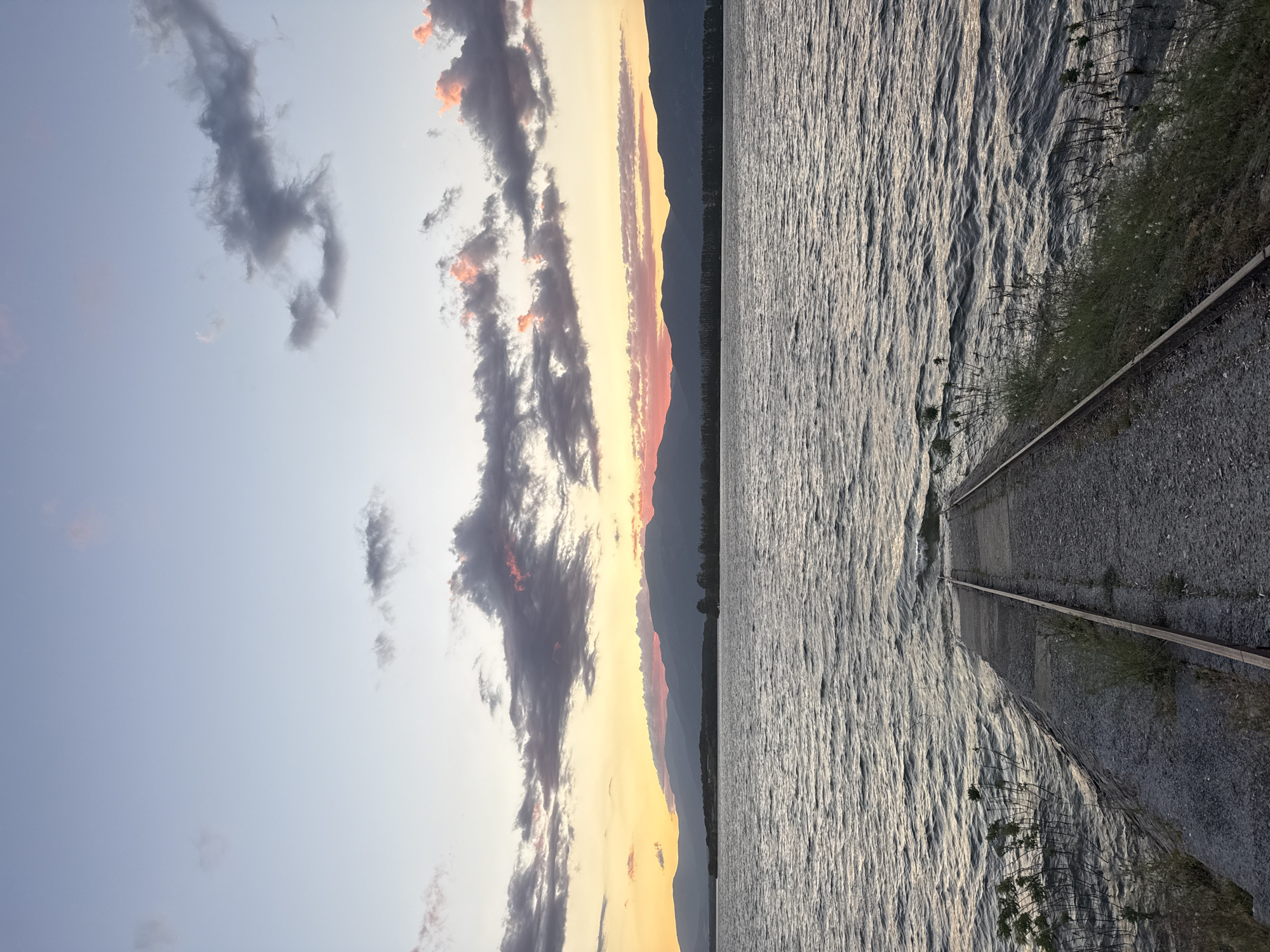 The image captures a serene sunset over Koprinka Reservoir in Koprinka, Bulgaria. In the foreground, a concrete boat ramp, lined with metal tracks, slopes gently into the rippling water. The water's surface reflects the soft hues of the twilight sky. To the left and right of the ramp, sparse green vegetation grows along the water's edge. In the background, a long, undulating mountain range is silhouetted against the horizon. The sky above is a canvas of soft blues, pinks, and oranges, with scattered clouds tinged by the setting sun. The overall scene evokes a sense of calm and natural beauty at the close of day.