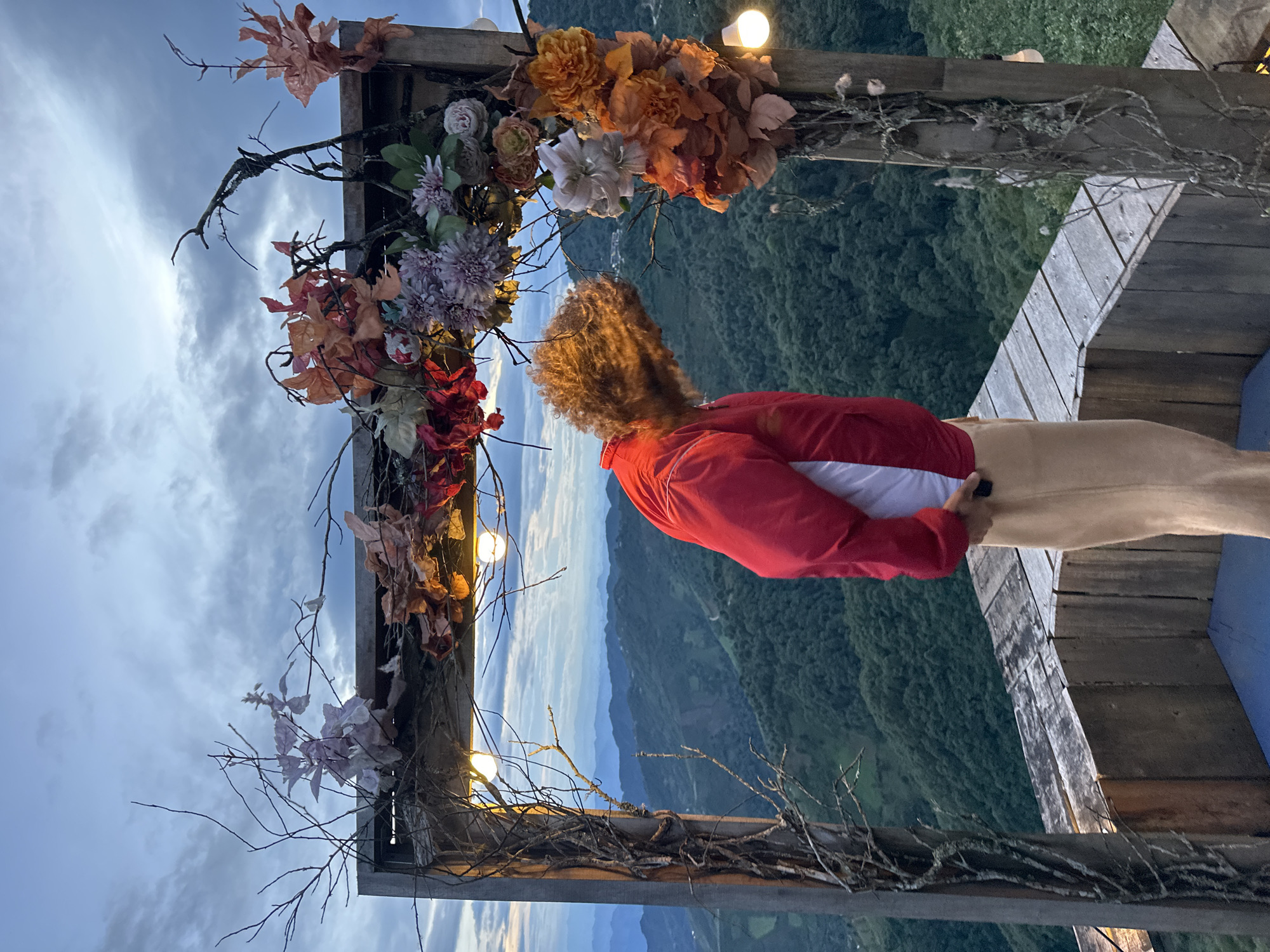 This image, captured in Ta Xua, Vietnam, presents an individual on a wooden viewing platform framed by a decorative structure, overlooking a vast mountainous landscape. The wooden frame, positioned centrally, is adorned with intertwined bare branches, several illuminated light bulbs along its top beam, and clusters of artificial flowers in shades of purple, red, orange, and white.
A person is situated slightly to the left of the frame's center, facing away from the viewer. They wear a red long-sleeved jacket over a white garment and light brown or beige pants. Their curly, reddish-brown hair appears in motion, largely obscuring their face as they lean forward and downwards. The individual's right hand is visible at their waistline, holding a dark, rectangular object.
Beyond the wooden structure, a panoramic view of multiple forested mountain ranges extends into the distance, with closer peaks displaying dense green foliage and more distant ranges appearing progressively lighter blue and hazy. The sky above is overcast with dense grey clouds, suggesting low light conditions or dusk. The wooden platform itself shows weathered planks, with a blue mat or surface visible on the ground directly beneath the person.