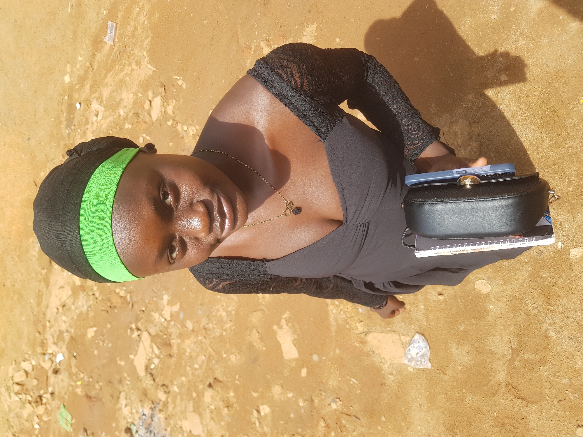 A young woman is captured from a high-angle perspective, outdoors, against a sandy, dirt ground. She is wearing a black dress with sheer, lace sleeves and a black headscarf with a bright green band. A gold necklace with a dark pendant rests on her décolletage. In her right hand, she holds a black, rounded purse, on top of which sits a blue-cased smartphone and a notebook. Her expression is one of direct gaze towards the camera. The sunlight casts a distinct shadow of the woman and her arm onto the ground, suggesting it is daytime. The ground appears to be somewhat uneven and scattered with small debris, some of which is white and reflective, possibly plastic. No visible text is present in the image.