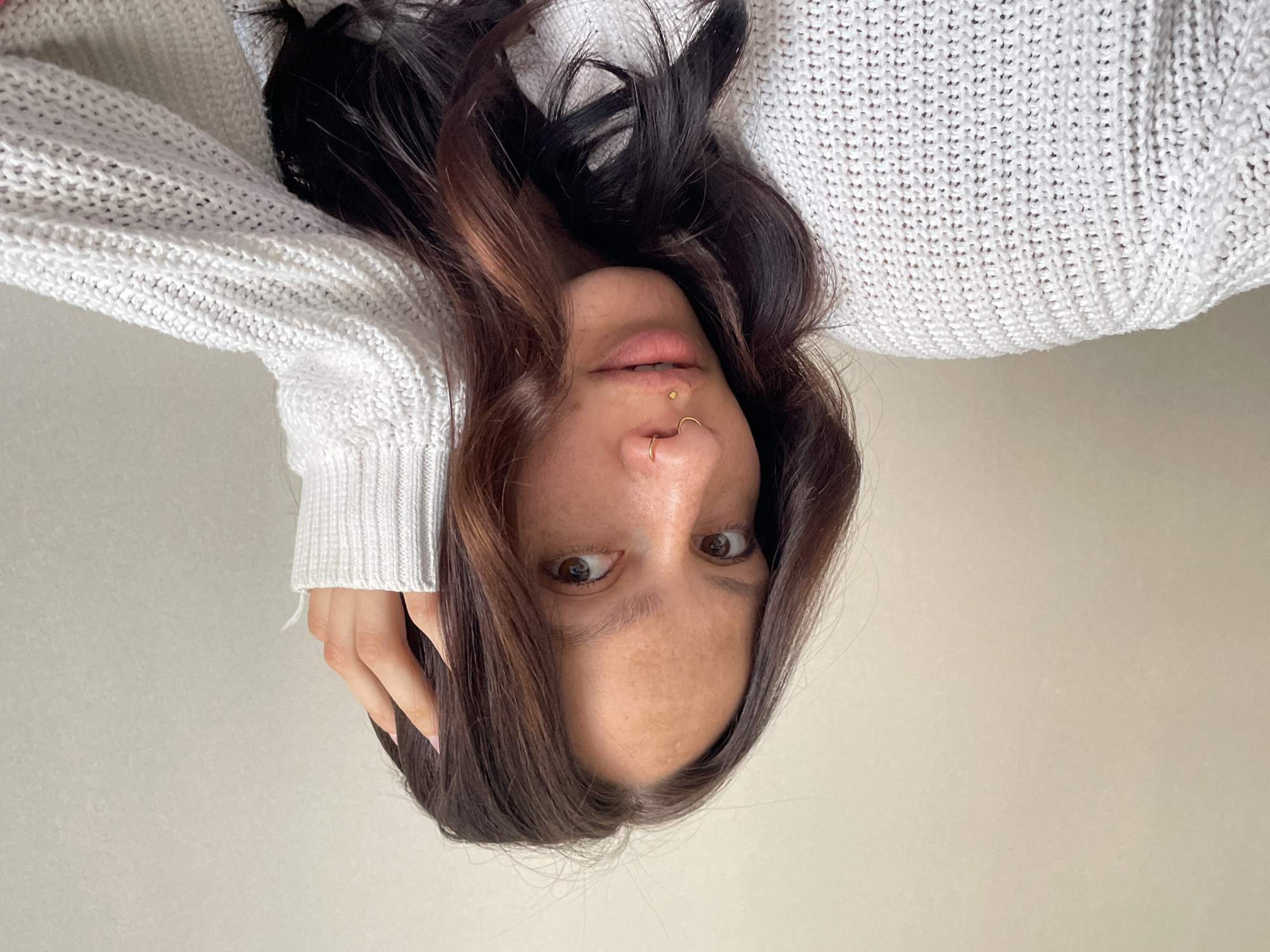 A woman with brown hair and facial piercings is lying down, wearing a white knit sweater. The background is a light beige wall. The location of the city of Izhevsk, Russia, is not ascertainable from the image.
