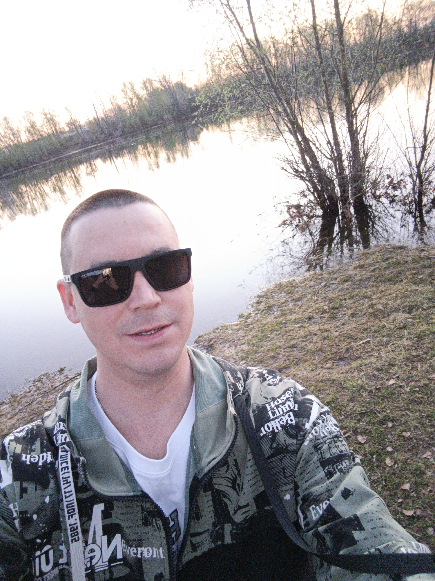 A man with a shaved head and sunglasses stands facing the camera. The reflection in his sunglasses shows someone holding a camera. He is wearing a light-colored t-shirt and a green, gray, and black printed jacket with a shoulder strap. Behind him is a body of water, possibly a river or lake, with trees lining the opposite bank. The water reflects the sky and the trees. The ground to the right of the man is covered in dry grass and sparse vegetation. The photo appears to have been taken near the water's edge in Chaikovsky, Russia. The environment suggests it is either late fall, winter, or early spring, based on the bare trees and dormant vegetation.