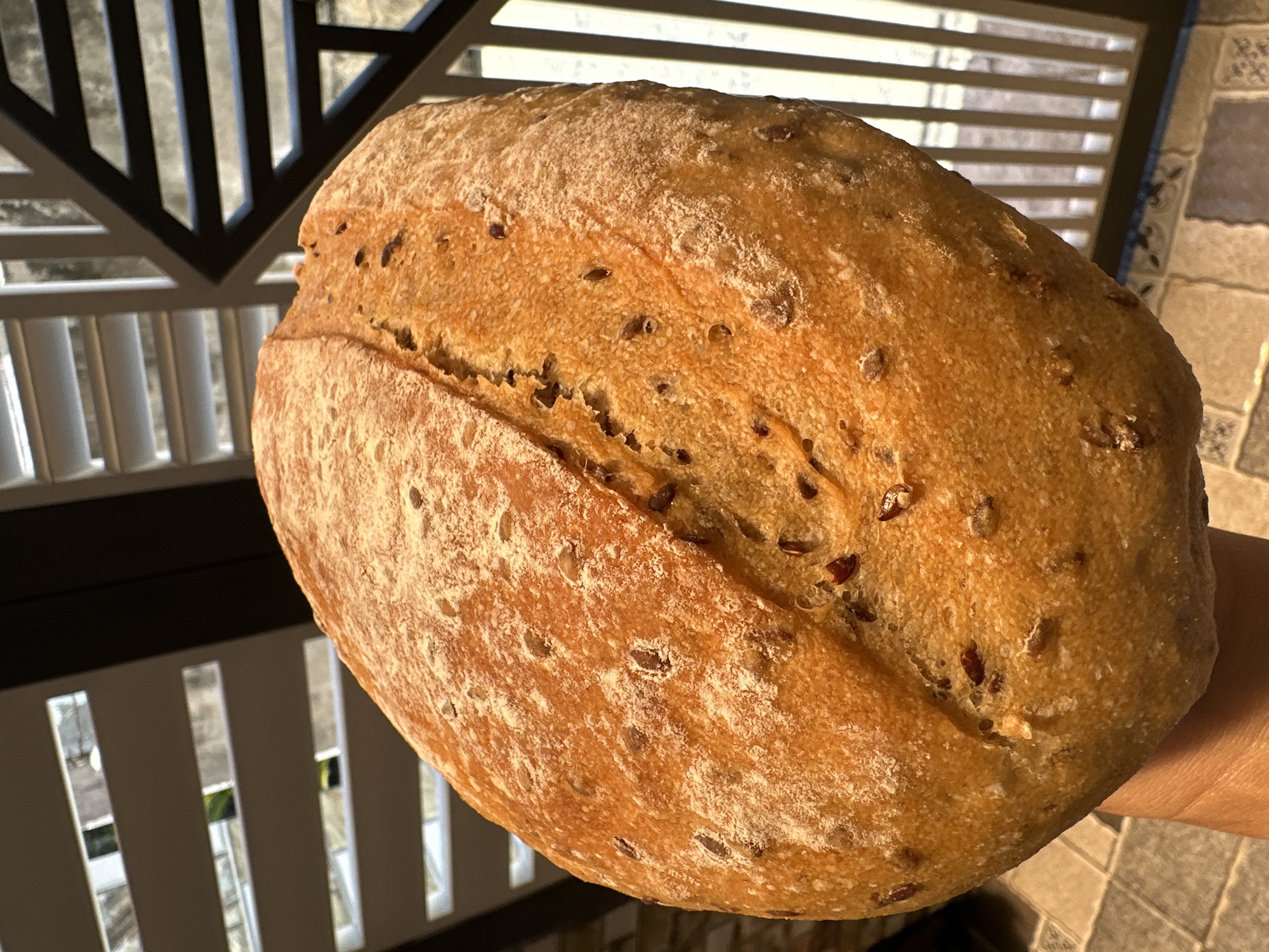 A close-up, well-lit image shows a loaf of seeded bread being held in someone's hand. The bread has a scored surface and is speckled with seeds. The background includes indoor architectural features, appearing to be a combination of decorative metalwork and blinds or slats. The scene is indoors and the location is determined to be Bac Nha Trang Ward, Vietnam.