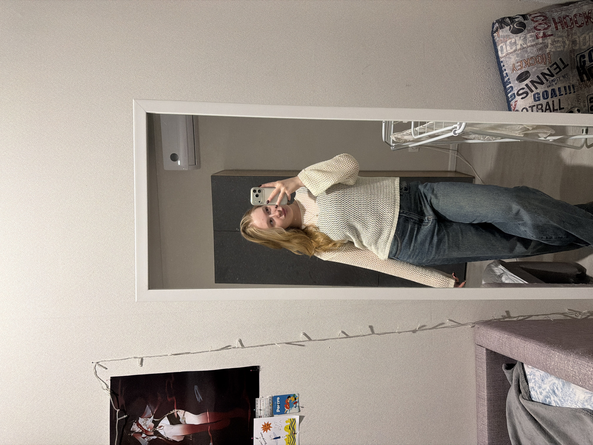 The image shows a person taking a selfie in a mirror. The person is wearing a light-colored sweater and jeans. In the background, there is a wall with a poster, some decorations, and a headboard. A shelf is also visible with items on it, including a cushion and what appears to be folded clothes. The location of Засечное, Russia, cannot be confirmed or denied based on the visual information in the image.