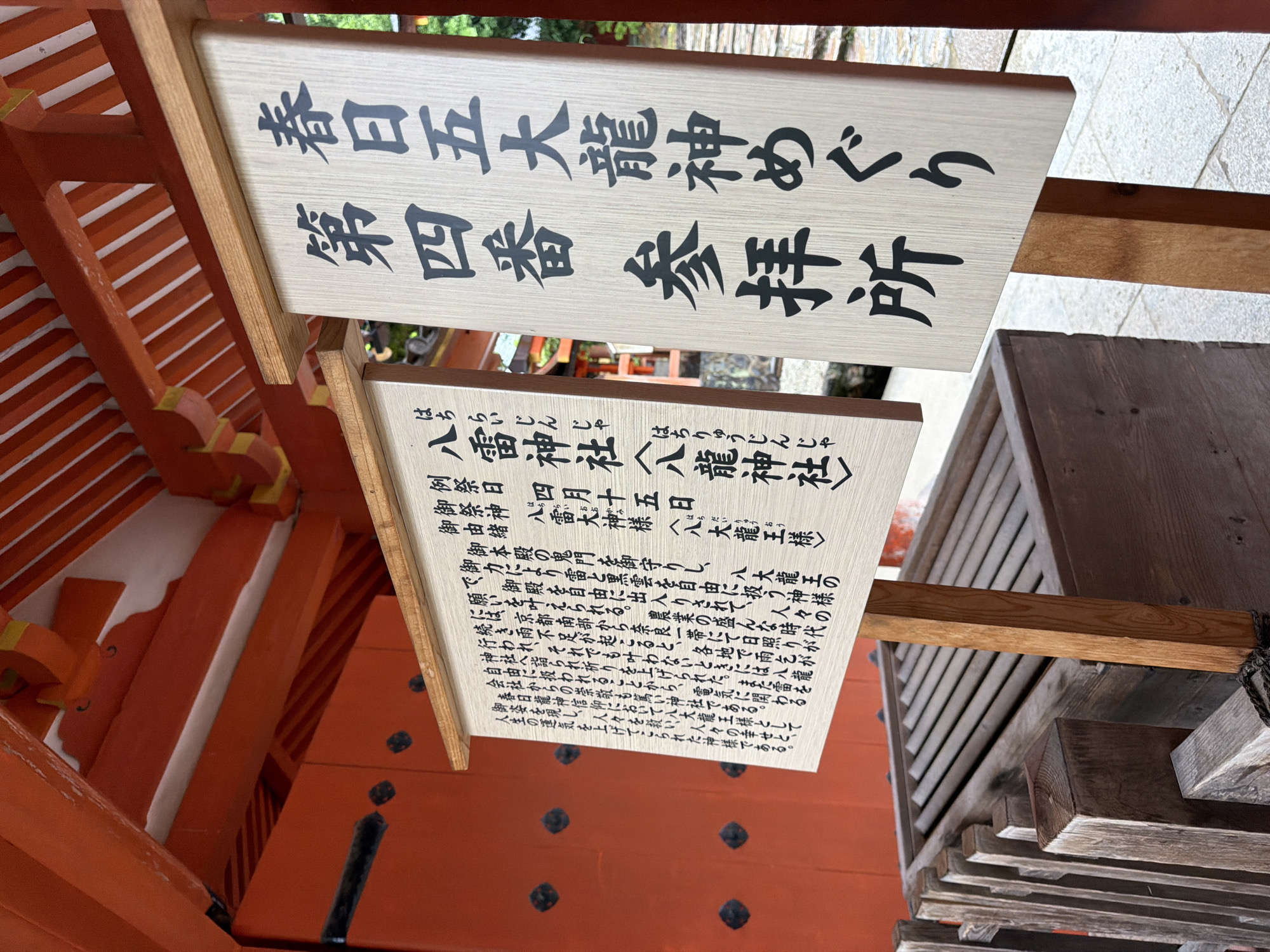 The image displays a detailed view of a worship area at the Hachirai Shrine (八雷神社), which is part of the Kasuga Taisha Shrine complex in Nara, Japan. Two prominent wooden signs with black Japanese characters stand in the foreground. The larger sign on the right identifies the location as the "Fourth Worship Place" (第四番 参拝所) for the "Kasuga Five Great Dragon Gods Pilgrimage" (春日五大龍神めぐり). The smaller sign on the left provides information about the Hachirai Shrine, identifying the enshrined deity as the "Eight Dragon God" (八龍神様), with smaller text detailing its origin and significance, including the regular festival date of April 15th. Behind the signs, vibrant orange-red traditional Japanese architectural elements, including structural beams and intricate ceiling components, are visible. In the lower left, a wooden offering box with horizontal slats on its lid is partially visible behind a low wooden railing, resting on a light grey, paved surface. The scene is devoid of people.