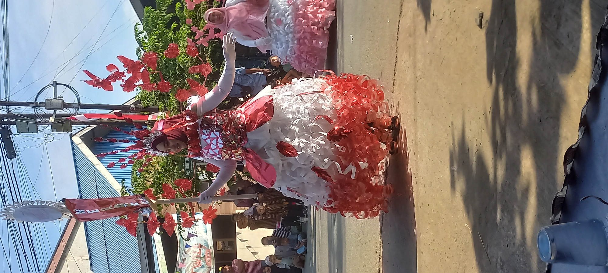 This image depicts an outdoor parade or festival scene in the city of Samarinda, Indonesia. The primary focus is on a young woman in a striking, elaborate costume. She wears a red hijab and a voluminous dress made of white and red, ruffled material, resembling layers of petals or ribbons. The costume is adorned with red floral accents and a silver, geometric-patterned bodice. She is holding a tall, decorative staff with a circular element at the top, also decorated with red floral elements. Her left arm is extended, and her right hand is also outstretched, suggesting she is interacting with or acknowledging a crowd.
To her right, partially visible, is another woman in a similar, though less elaborate, dress, this one predominantly pink and white. Both women appear to be participating in a performance or parade.
The background reveals a street lined with buildings and trees under a clear blue sky. A crowd of onlookers, including children and adults, are seated or standing on the side of the street, watching the parade. Some individuals are holding phones, likely recording the event. Utility poles with wires are visible in the background, a common feature in urban Indonesian settings. The ground is paved with asphalt. A banner with text is visible on the left side of the image, but it is partially obscured and difficult to read. The overall atmosphere suggests a celebratory public event.