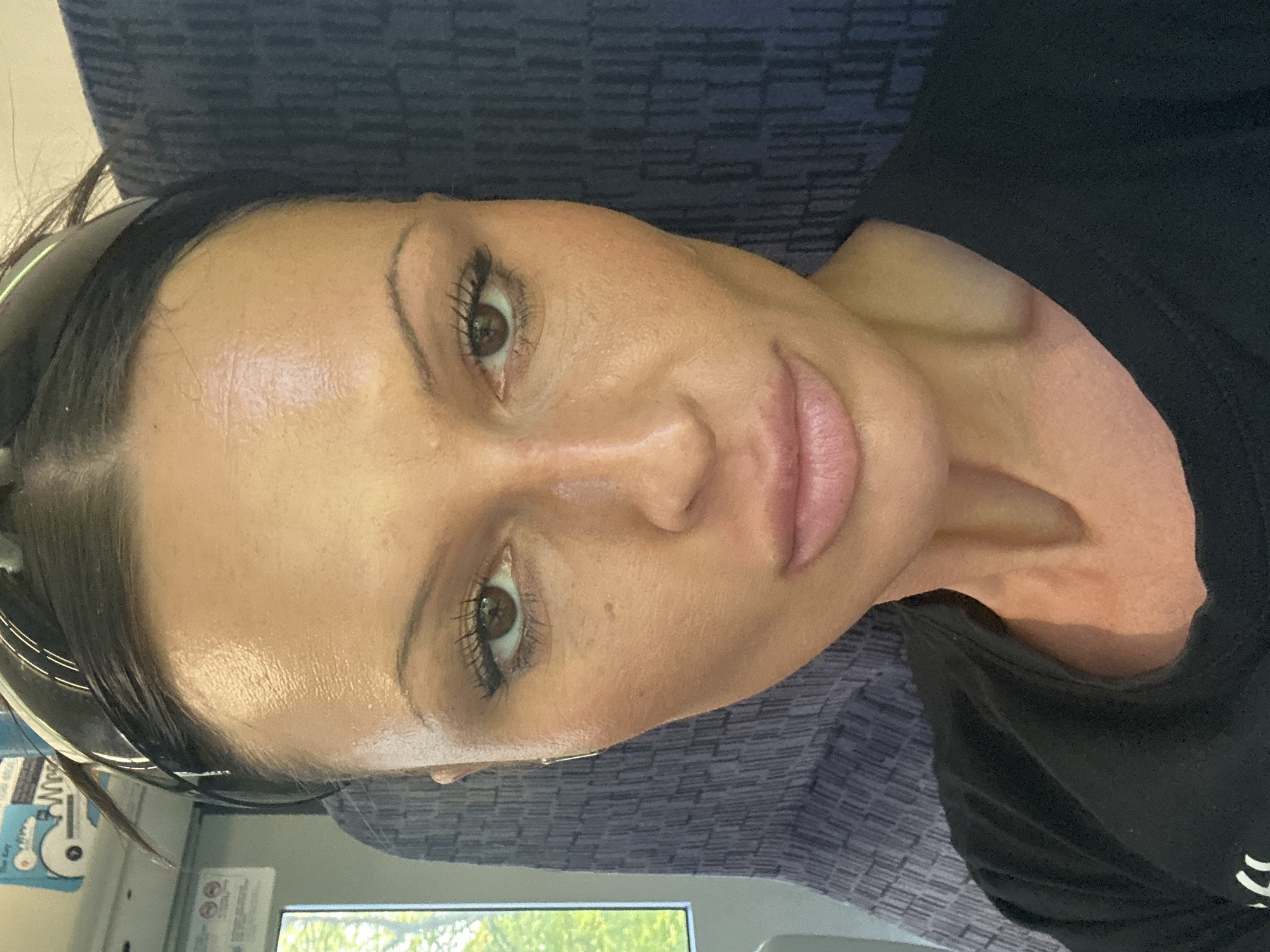 A woman is seated in what appears to be a train. The lighting is bright. The woman has dark brown hair, brown eyes, and light skin. She is wearing a black headband and a black t-shirt. The seat is upholstered with a blue patterned fabric. A window is visible, showing green foliage. The location appears to be Reigate and Banstead, United Kingdom.
