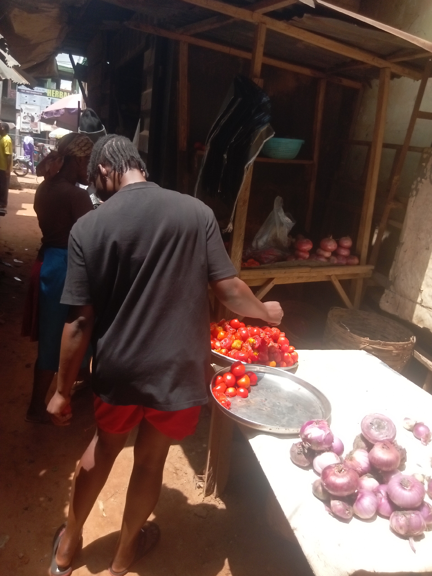 The image shows an outdoor scene in Ogidi, Nigeria. In the foreground, a person wearing a black t-shirt and red shorts is selecting tomatoes from a silver tray. The tray is on a white table which also holds a pile of red onions. Behind the person is another person wearing a headwrap and a blue skirt. Further back, other people are visible in the background on a dusty path. A wooden structure with shelves is behind the table, displaying more tomatoes and other items. The lighting suggests a sunny day.