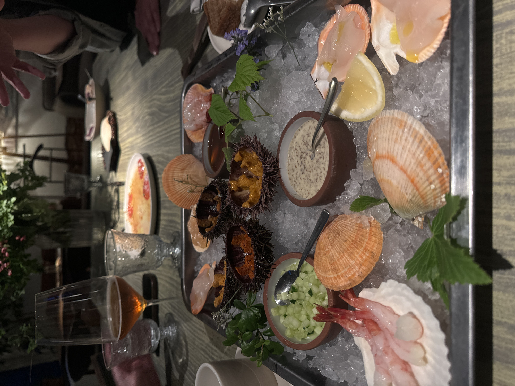 This image depicts a seafood platter served on a bed of ice, presented on a metal tray. The platter contains several types of seafood, including sea urchins, scallops, and shrimp. Accompaniments include small bowls of what appears to be chopped cucumber and a creamy sauce, possibly a dressing or aioli. A glass of amber-colored liquid, likely wine or beer, is visible to the left, along with other drinking glasses and a person's hand in the background. The setting appears to be a restaurant, with a wooden table surface and a blurred background suggesting an indoor environment. The lighting is dim, creating a mood typical of an evening dining experience. The image does not provide any specific visual cues to identify the location as Saint Petersburg, Russia.