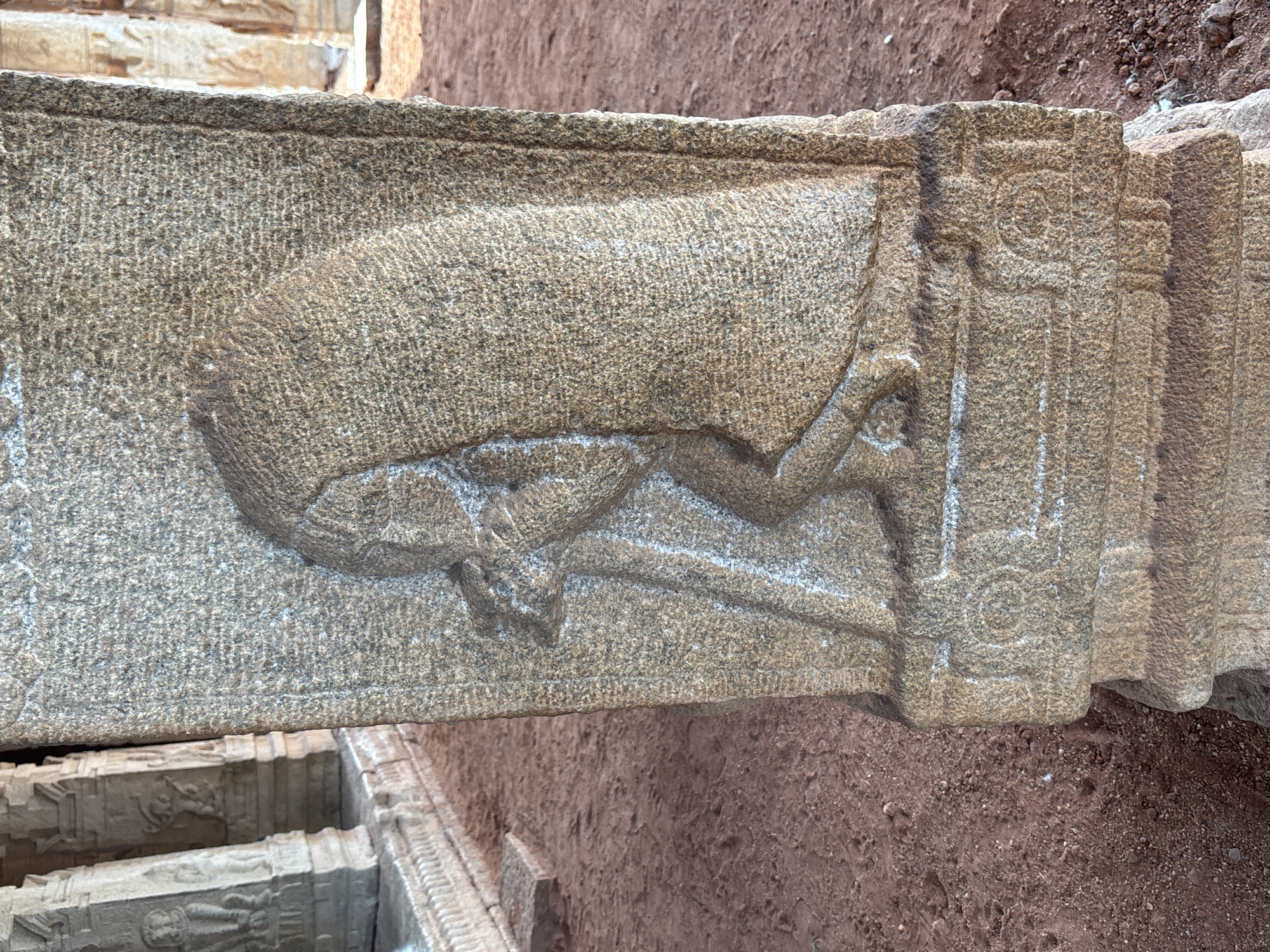 A close-up, upward-tilted shot captures a section of a weathered stone structure featuring a bas-relief carving, presumably located in or near Kamalapura, India. The carving depicts a horizontal, stylized figure, possibly an animal or mythical creature, sculpted into the stone's surface. The subject's posture suggests a reclining or falling position. The stone is textured with a coarse, grainy surface, indicating its age and exposure to the elements. To the right of the carving, the stone is fashioned into decorative columns or pilasters with carved linear and geometric motifs. The background is predominantly reddish-brown soil.