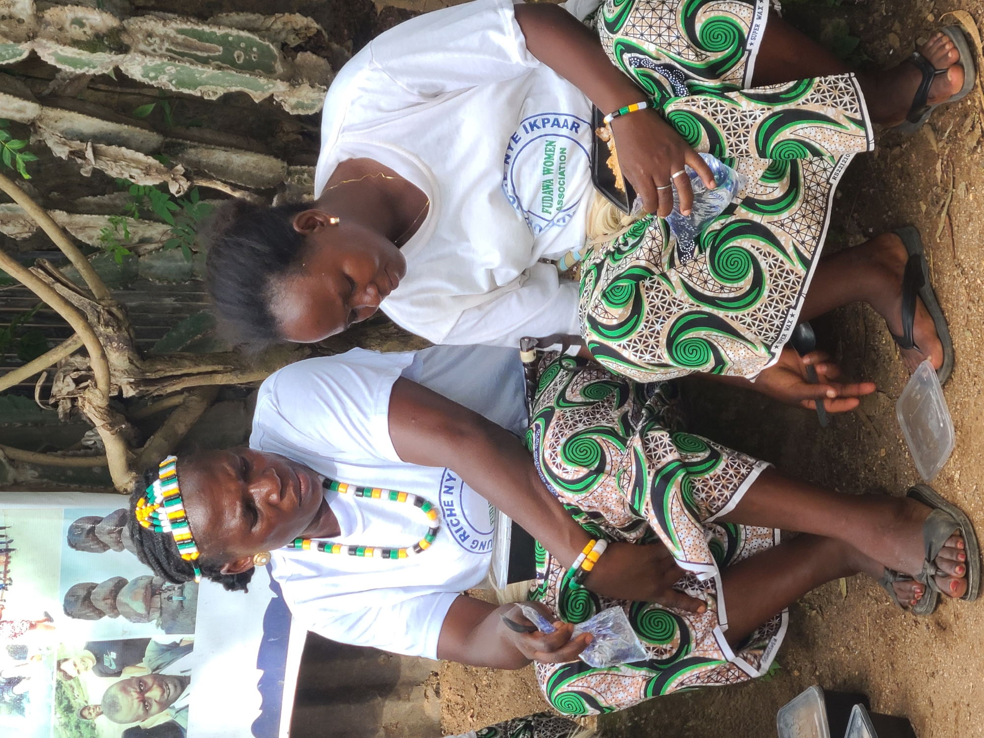 Two women are seated outdoors in a sandy area, possibly in Jos, Nigeria. The woman on the left, wearing a white t-shirt and a colorful beaded headband and necklace, is looking down. She has a plastic bag with a blue object inside in her left hand. The woman on the right, also wearing a white t-shirt, is looking down and holding a black food container in her right hand. Both women are wearing vibrant, patterned skirts with green, brown, and white swirls. They are barefoot. In the background, there is a backdrop with images and text, and behind that, large, textured tree trunks or cacti. Several black and clear plastic food containers are scattered on the ground around them.
